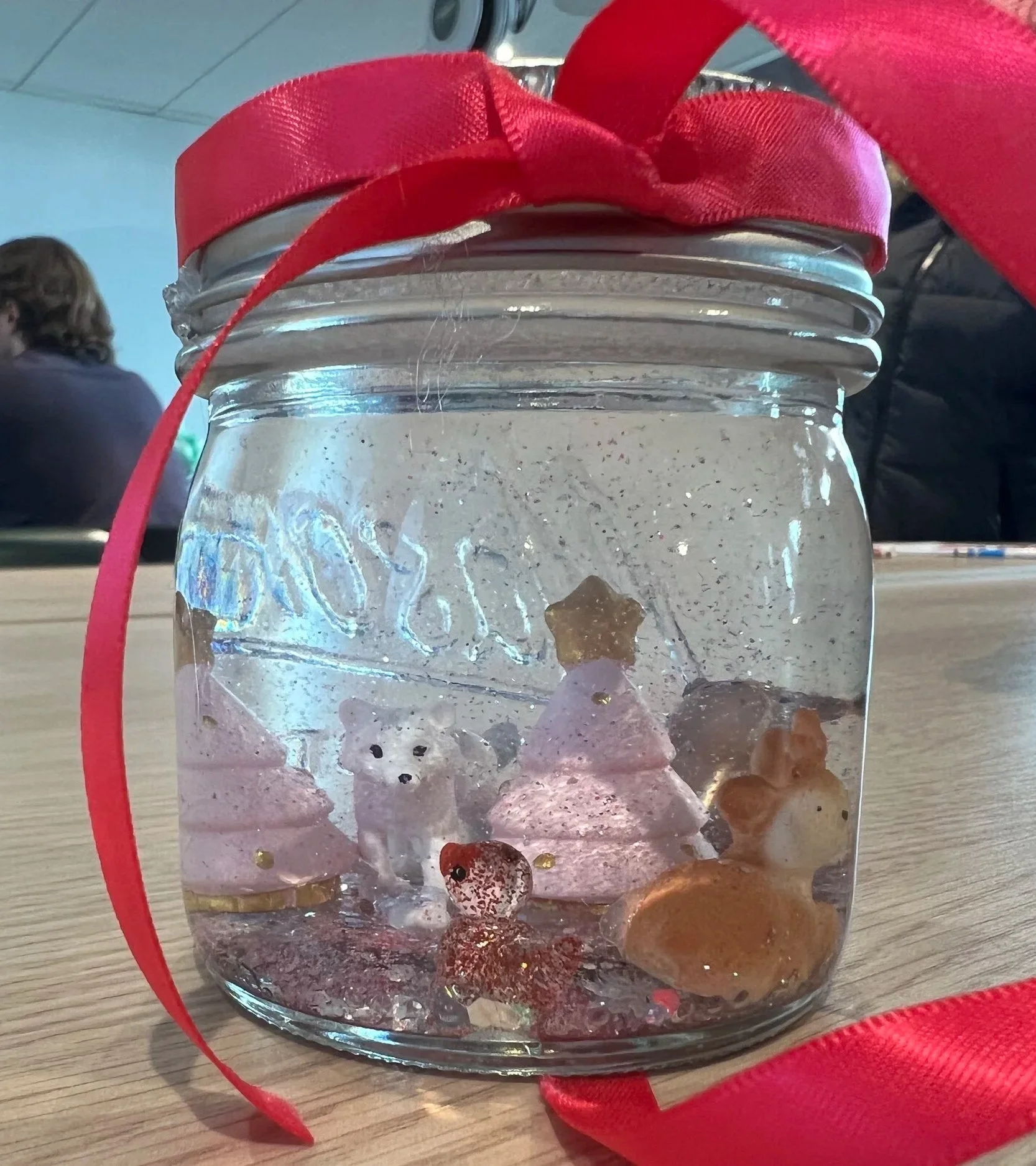 A handmade snow globe made from a mason jar, with small holiday decorations inside and a red ribbon at the top.