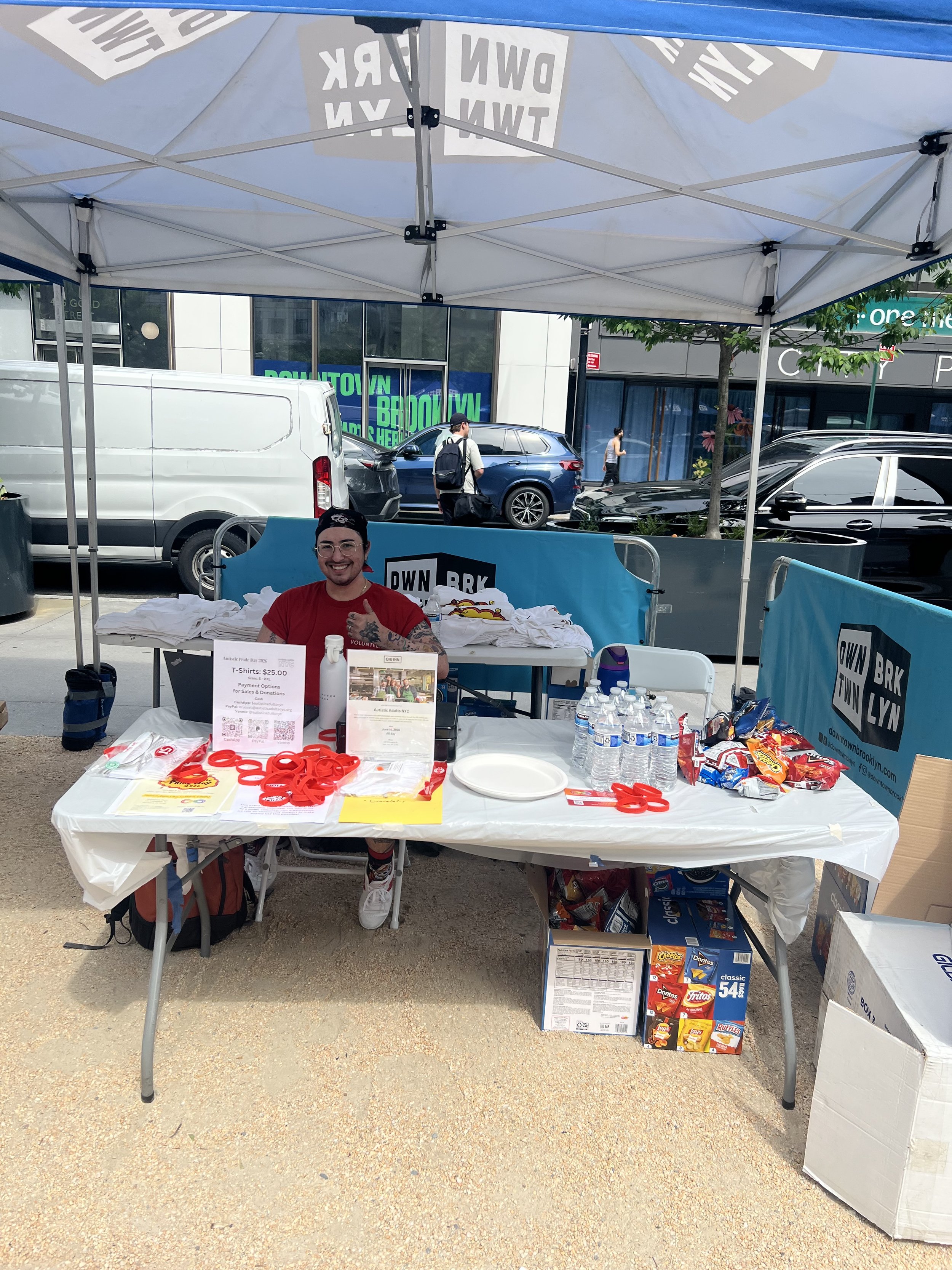  Table with Autistic Pride Day merchandise. Behind the table an AANYC volunteer is giving a thumbs up 