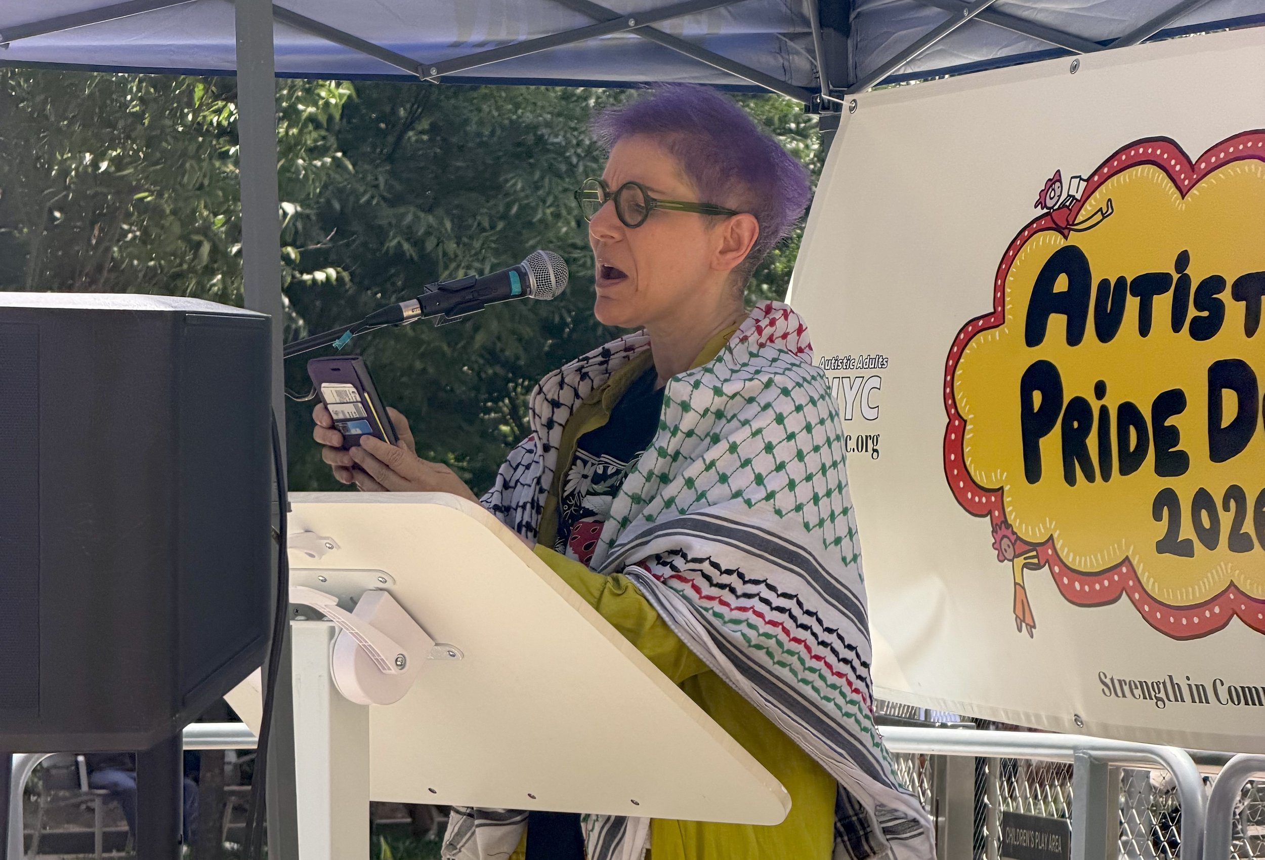  Person with purple hair and a keffiyeh speaking on stage 