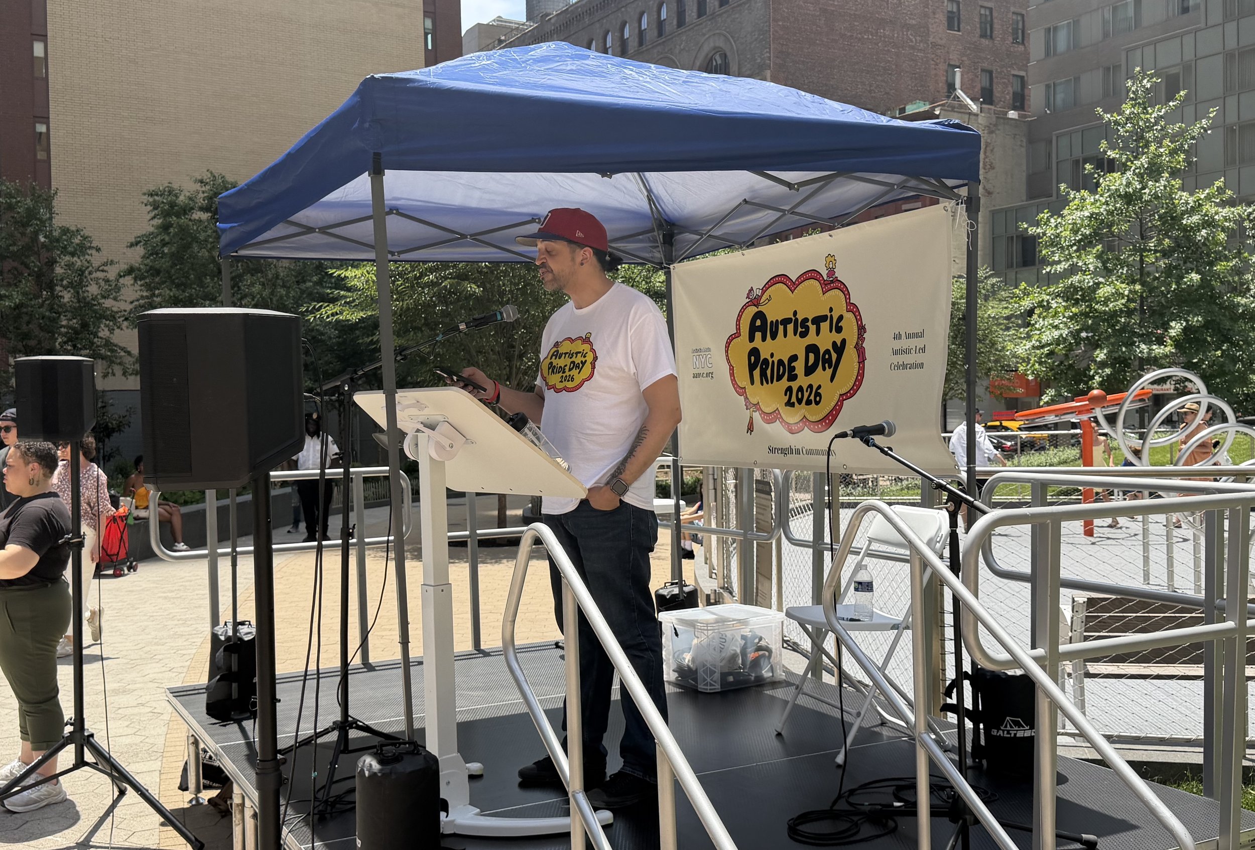  Person speaking on stage with a white “Autistic Pride Day 2026” shirt and a red hat 