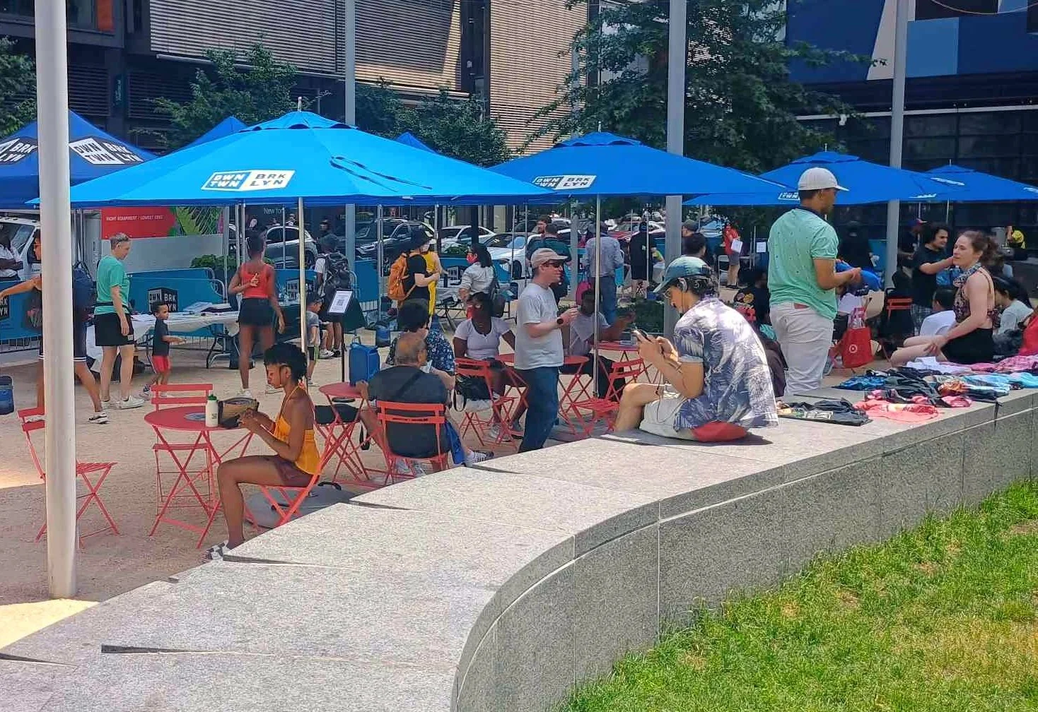  Crowd underneath blue umbrellas above picnic tables. Shirts are lined up along a concrete wall 