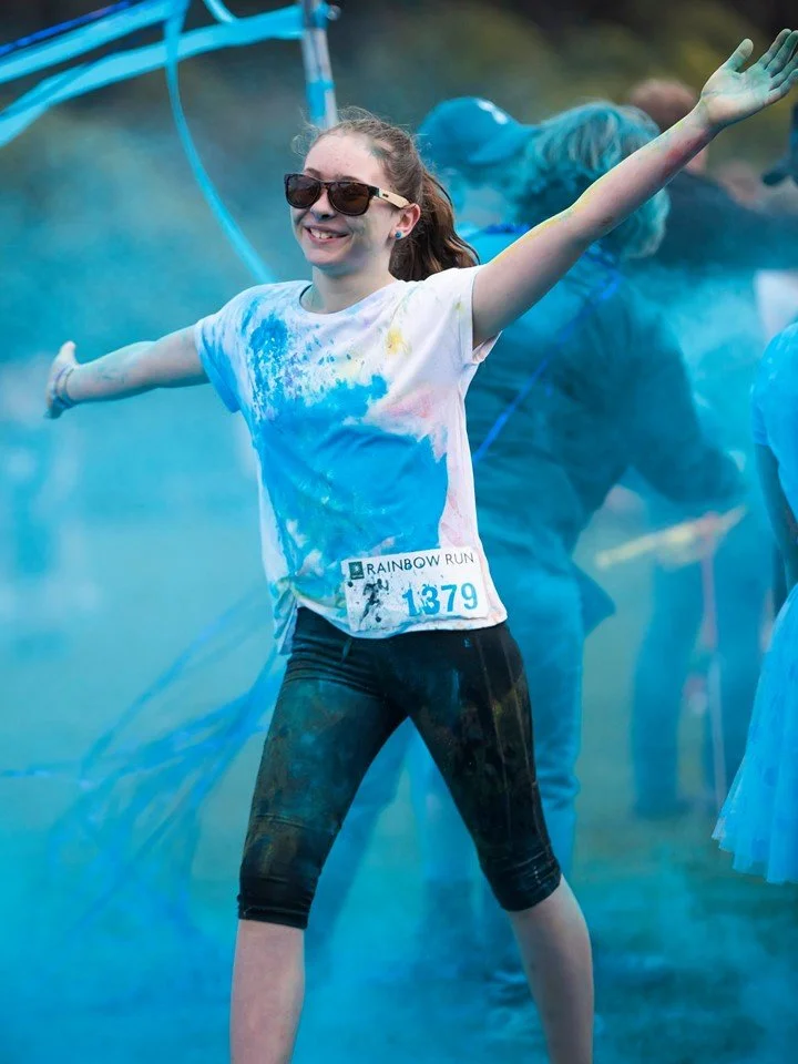 Young girl with sunglasses and a white T-shirt covered in blue powder, smiling with arms outstretched at a color run event, surrounded by blue powder and other participants.