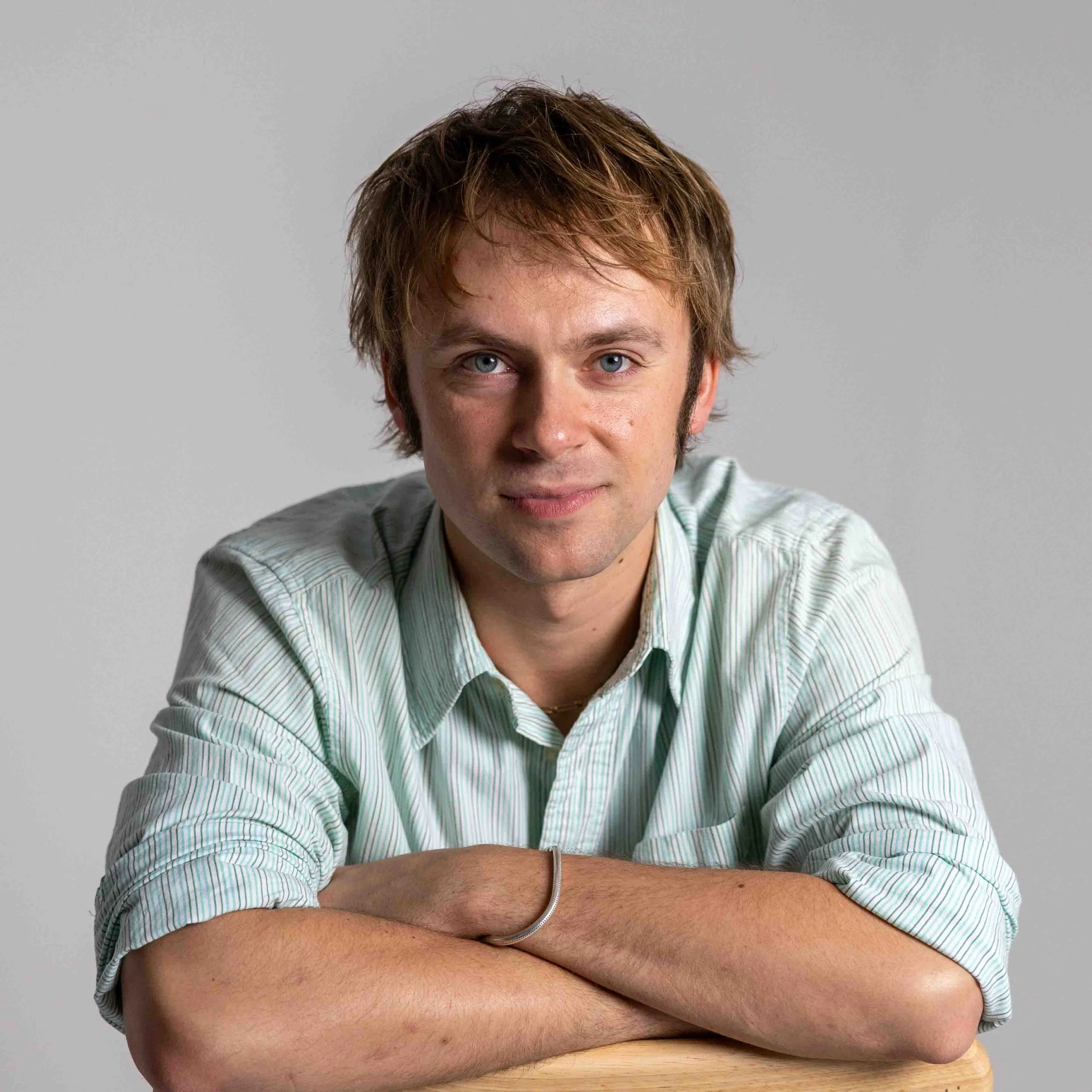 Portrait of a young man with light brown hair, blue eyes, wearing a light green striped shirt, sitting with arms crossed, against a plain gray background.