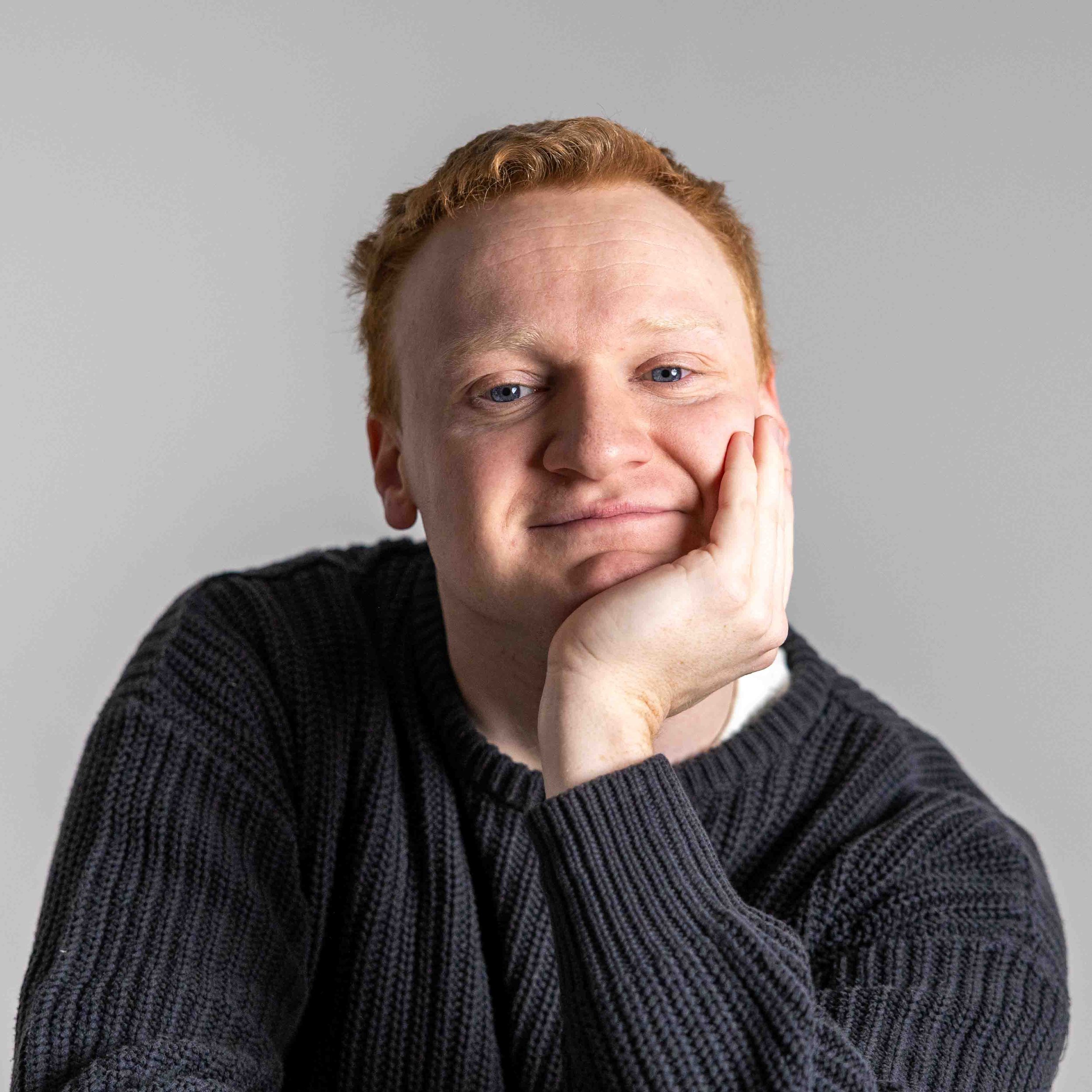 A young man with ginger hair and blue eyes resting his chin on his hand, wearing a black ribbed sweater, against a plain grey background.