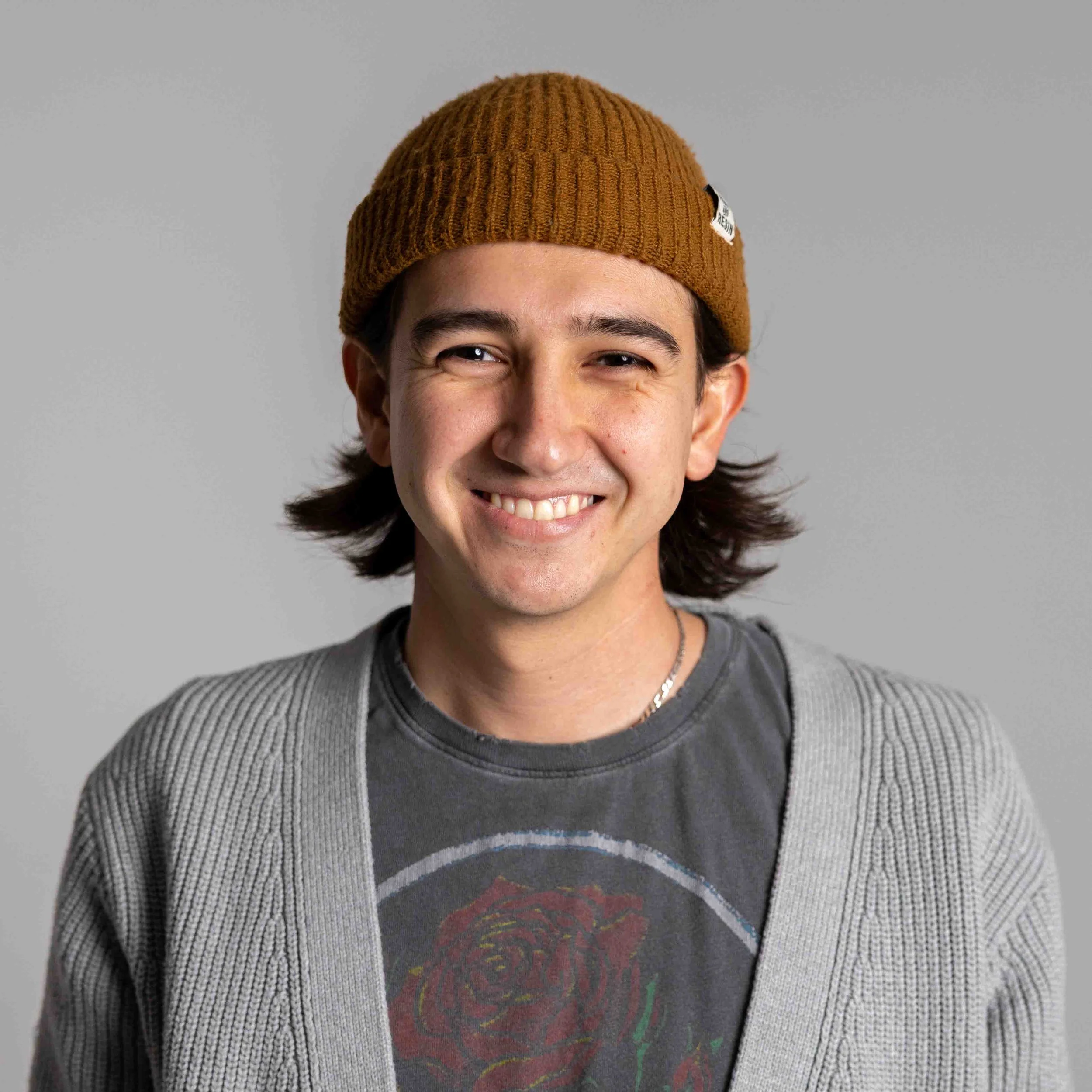 A young man with medium-length dark hair wearing a brown knitted beanie hat, a gray cardigan, and a black t-shirt with a red rose graphic, smiling at the camera against a plain gray background.