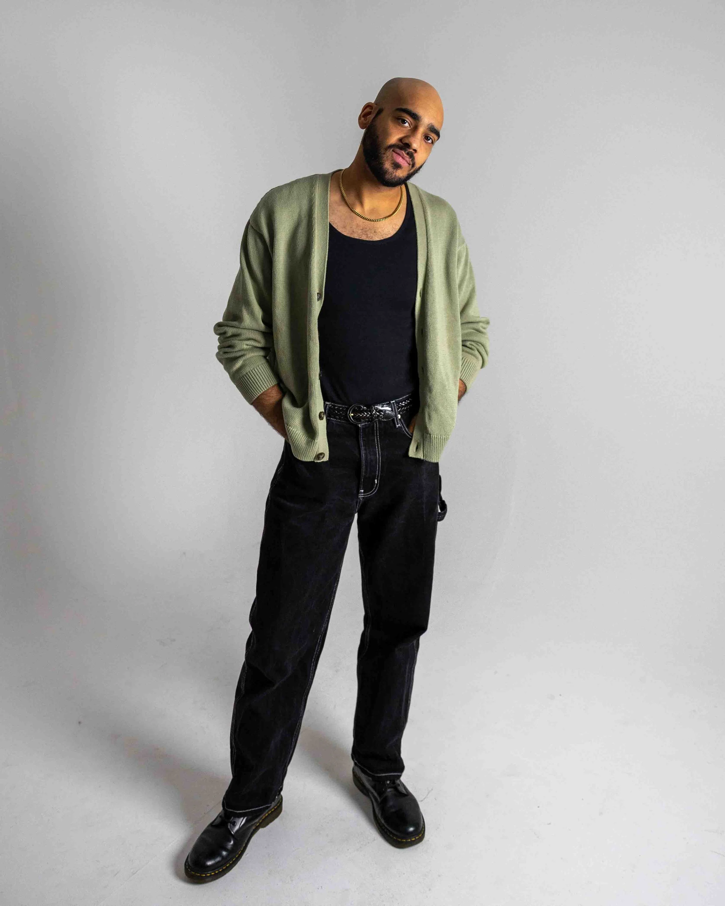 A man with a shaved head, beard, and mustache stands with hands in pockets, wearing a black t-shirt, green cardigan, black jeans, and black boots, against a plain light gray background.