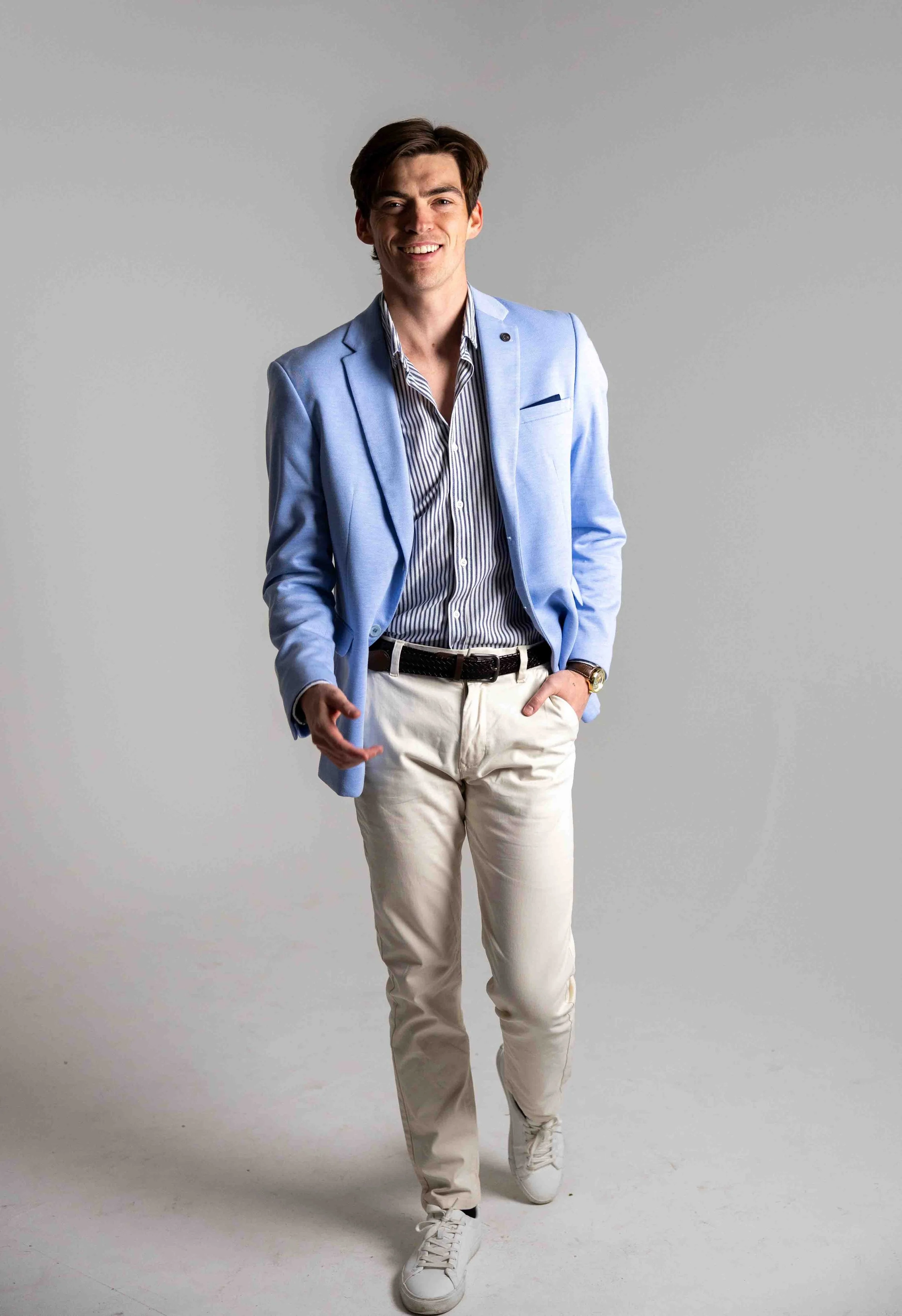 A man smiling and walking towards the camera wearing a light blue blazer, striped shirt, beige pants, and white sneakers, with a gray background.