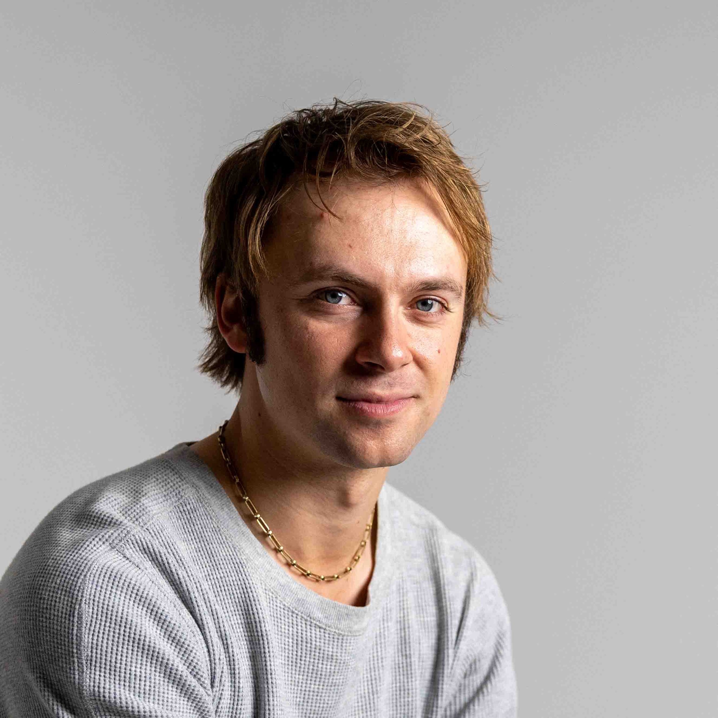 Portrait of a young man with red hair and blue eyes wearing a gray shirt and gold chain necklace.