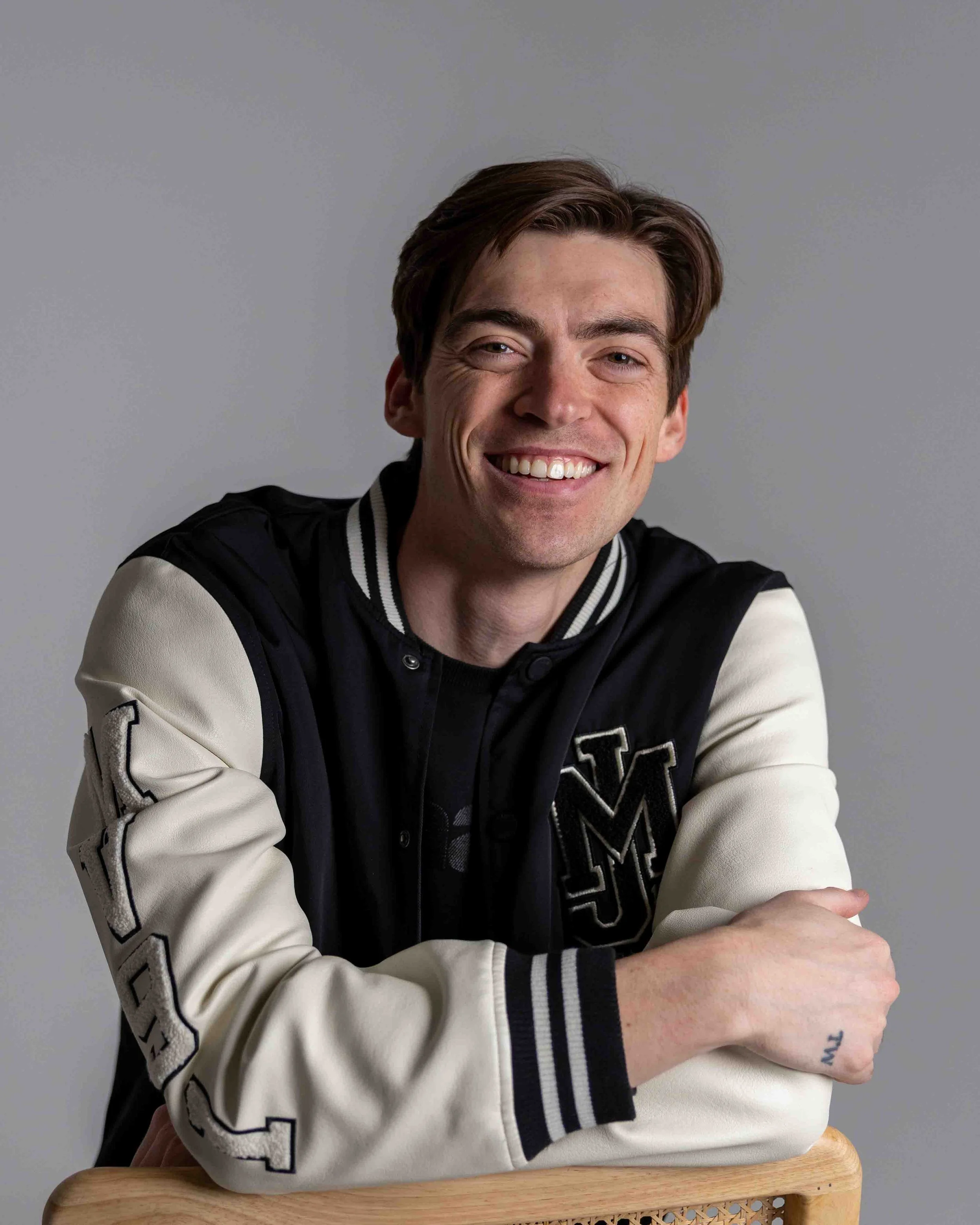 A young man with brown hair and fair skin, smiling at the camera, wearing a black and white varsity jacket with letter patches, sitting with his arms crossed on the back of a wooden chair, against a plain gray background.