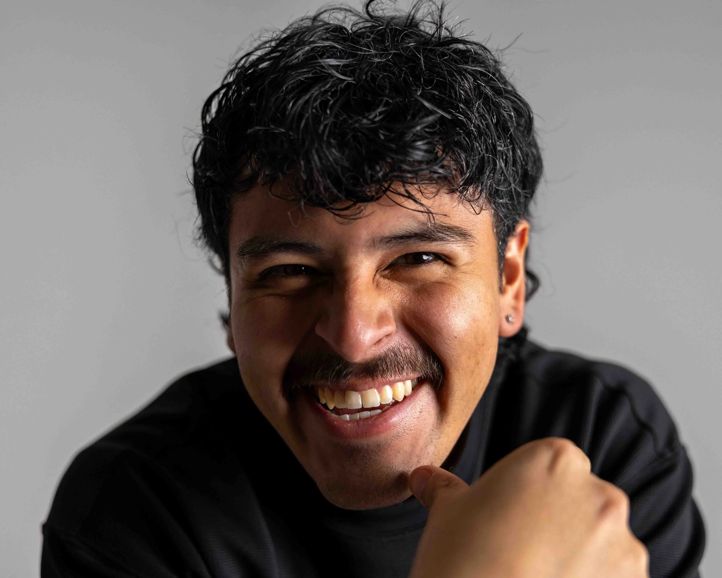 A man with dark curly hair, a mustache, and earrings smiling broadly, with his right fist raised in front of his face, against a plain gray background.