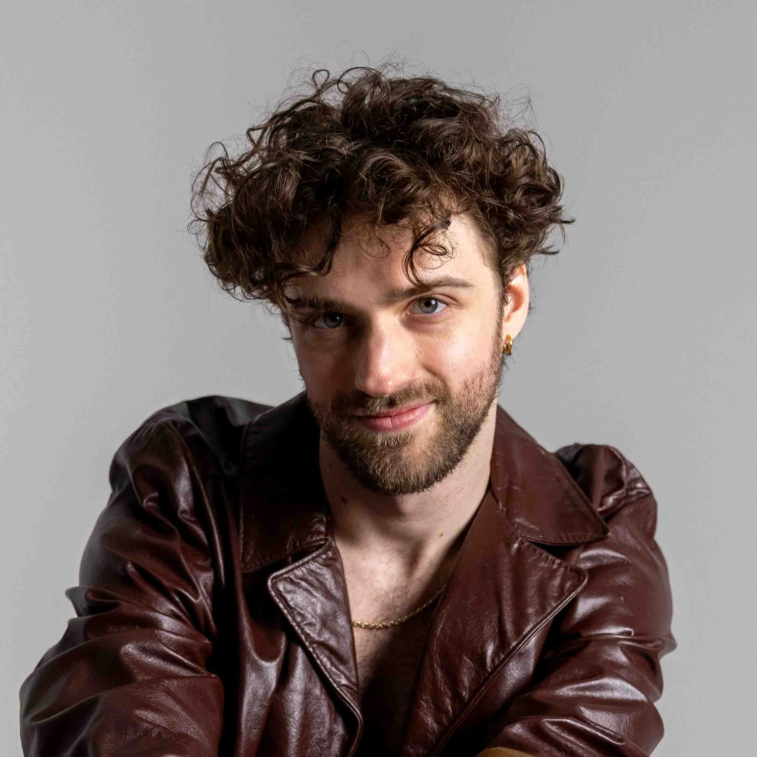 A young man with curly brown hair, beard, and blue eyes wearing a brown leather jacket, gold chain, and gold earring, looking at the camera against a neutral background.