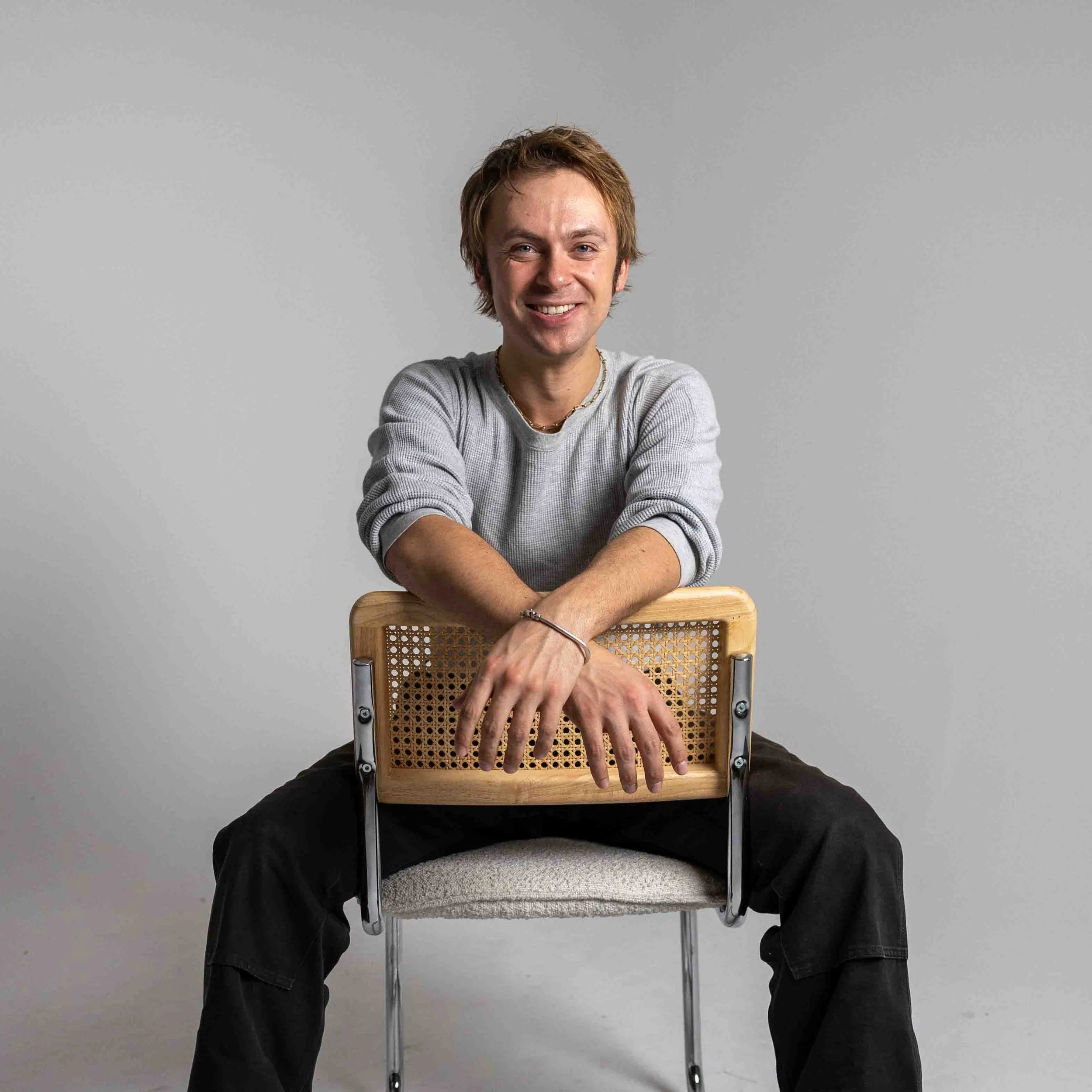 A young man with light brown hair, wearing a gray long-sleeve shirt and black pants, sitting on a chair with a woven backrest, smiling at the camera against a plain gray background.