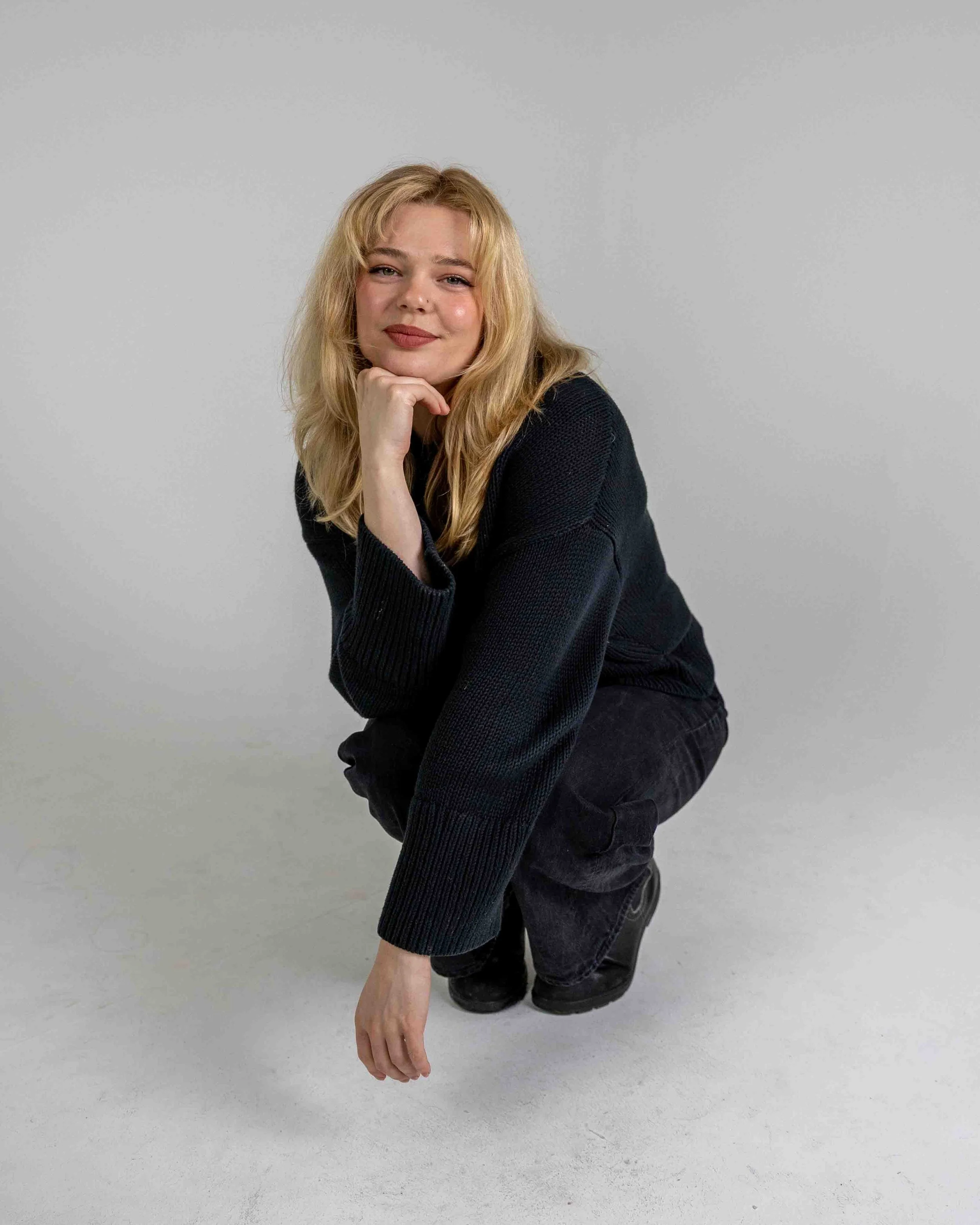 A young woman with blonde hair, wearing a black sweater and black pants, kneeling on the ground with her right hand touching her chin, smiling softly at the camera against a plain gray background.
