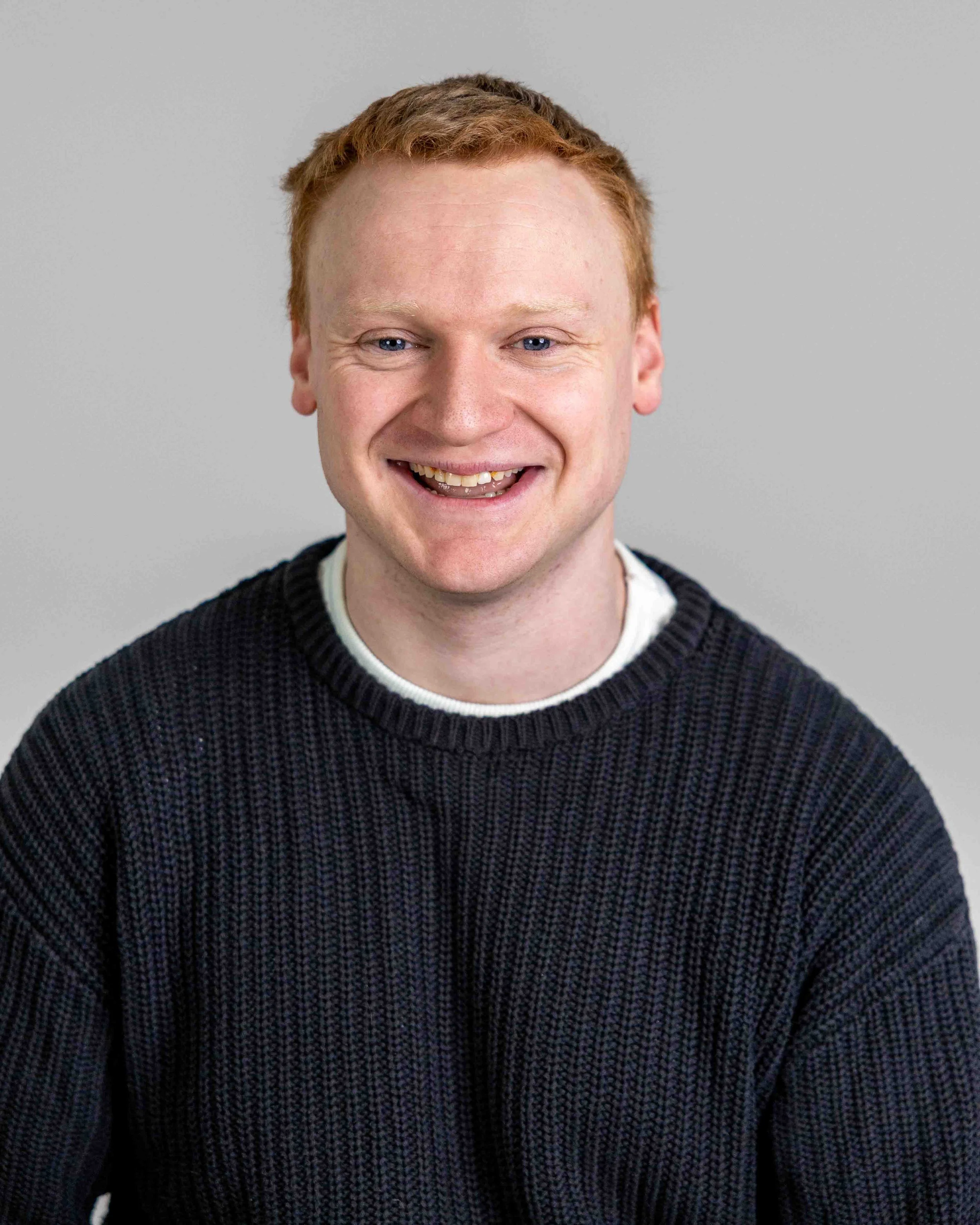 A man with red hair and a big smile wearing a black sweater over a white shirt, posing in front of a neutral gray background.