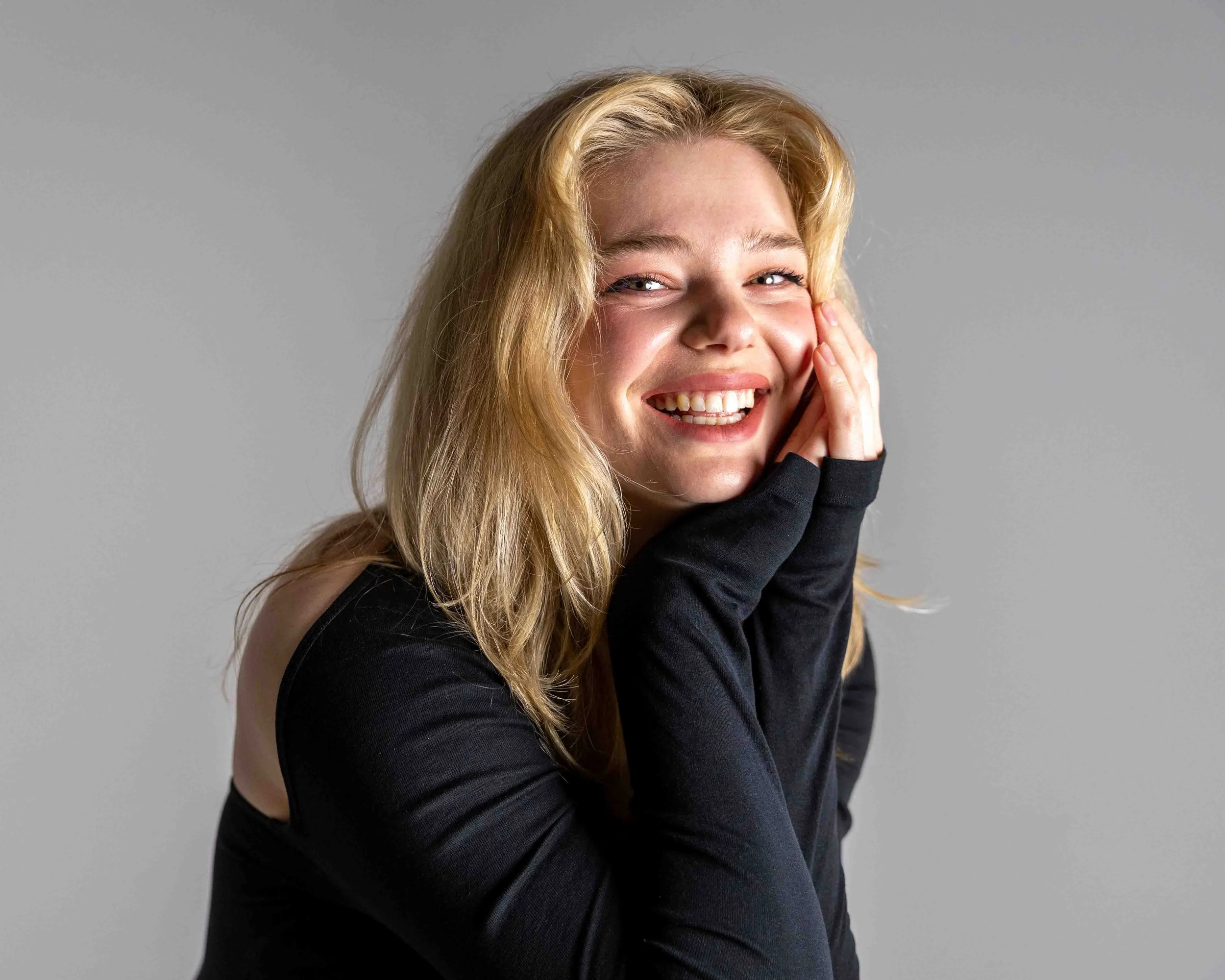 A young woman with long blonde hair smiling and holding her hand to her cheek, wearing a black top against a gray background.