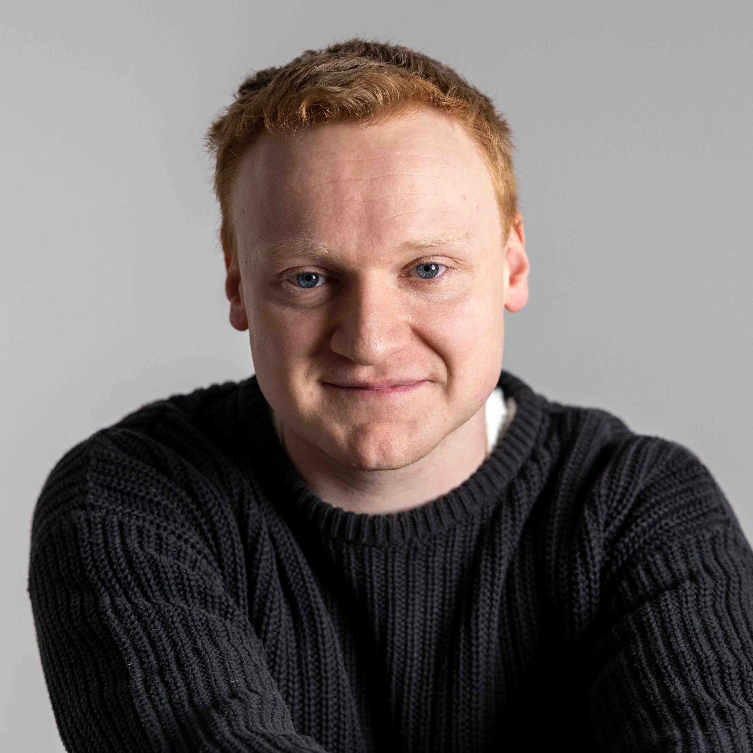 Portrait of a young man with red hair and blue eyes, wearing a black knitted sweater, smiling softly against a plain gray background.