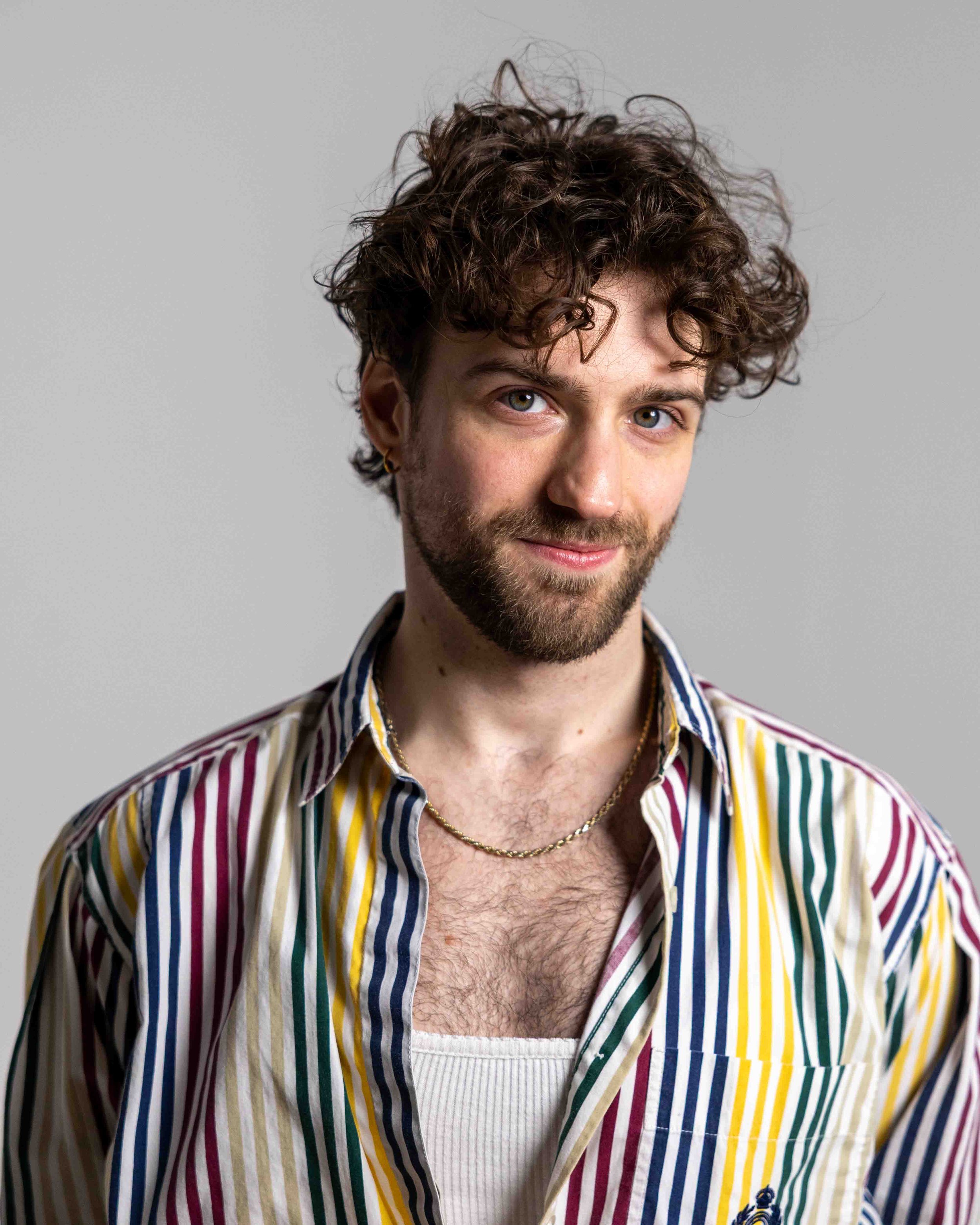 A young man with curly brown hair, beard, and blue eyes, smiling slightly, wearing a striped multicolored shirt over a white tank top, accessorized with a gold chain necklace and small hoop earring, against a plain gray background.