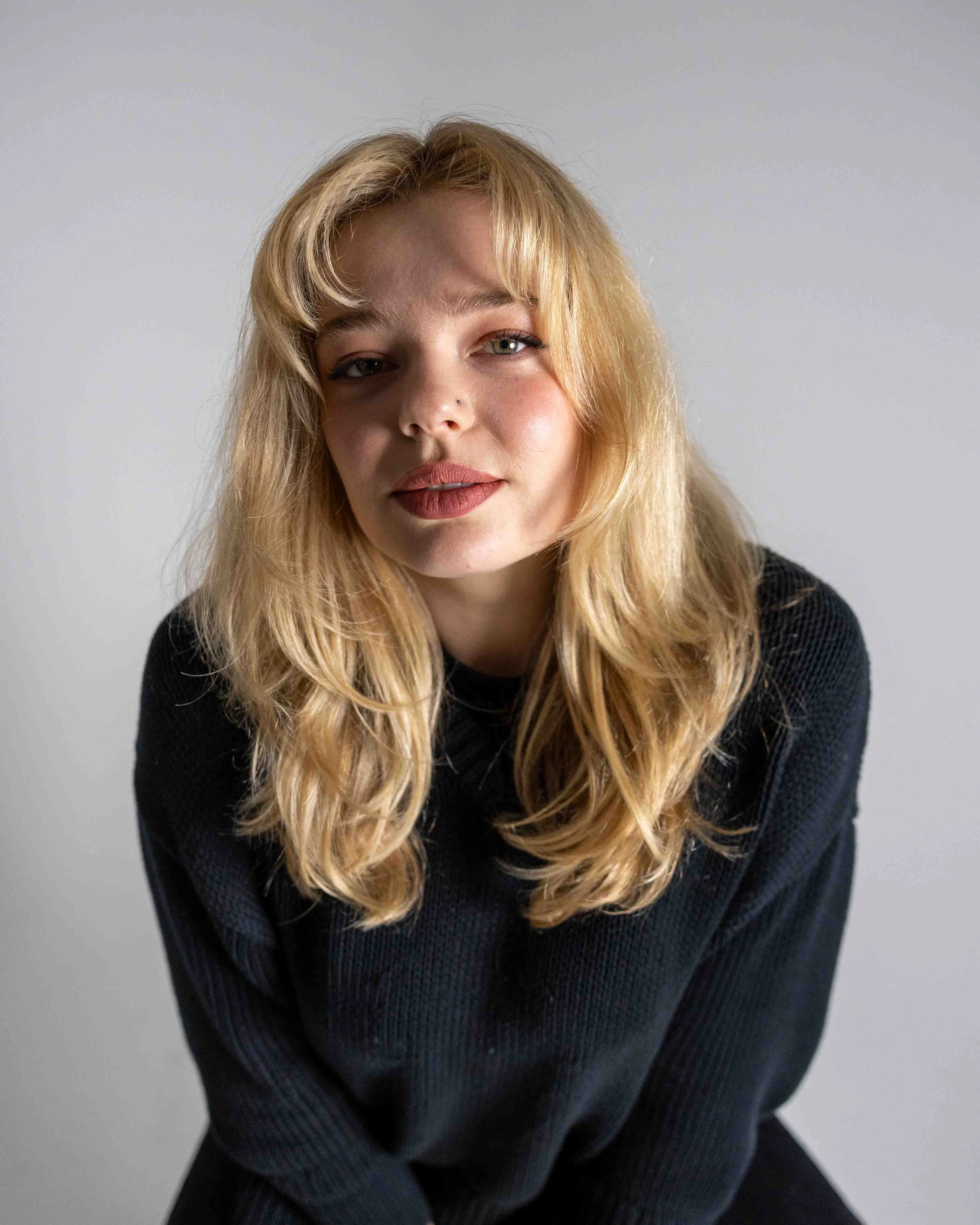 A woman with long blonde hair wearing a black sweater, looking directly at the camera against a plain white background.