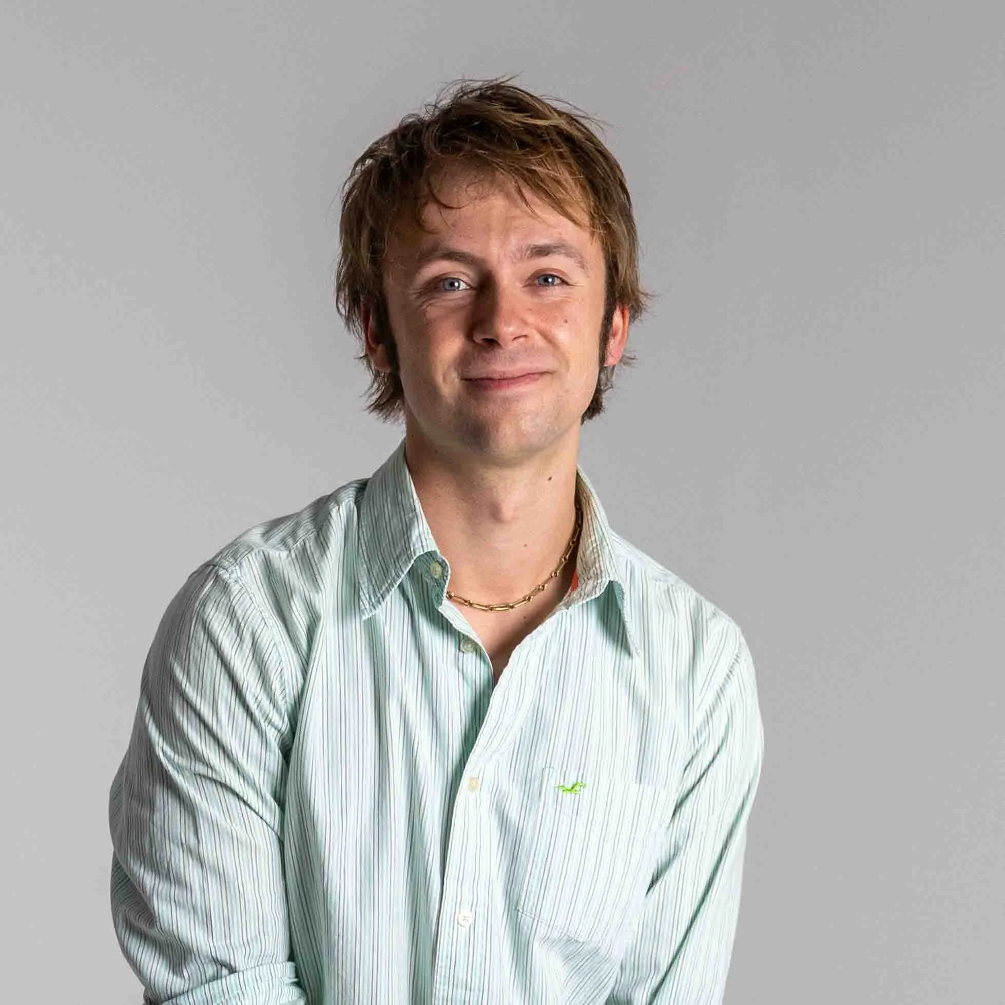 A young man with messy brown hair, blue eyes, and fair skin wearing a light green striped button-up shirt and a gold chain necklace, smiling slightly against a plain gray background.