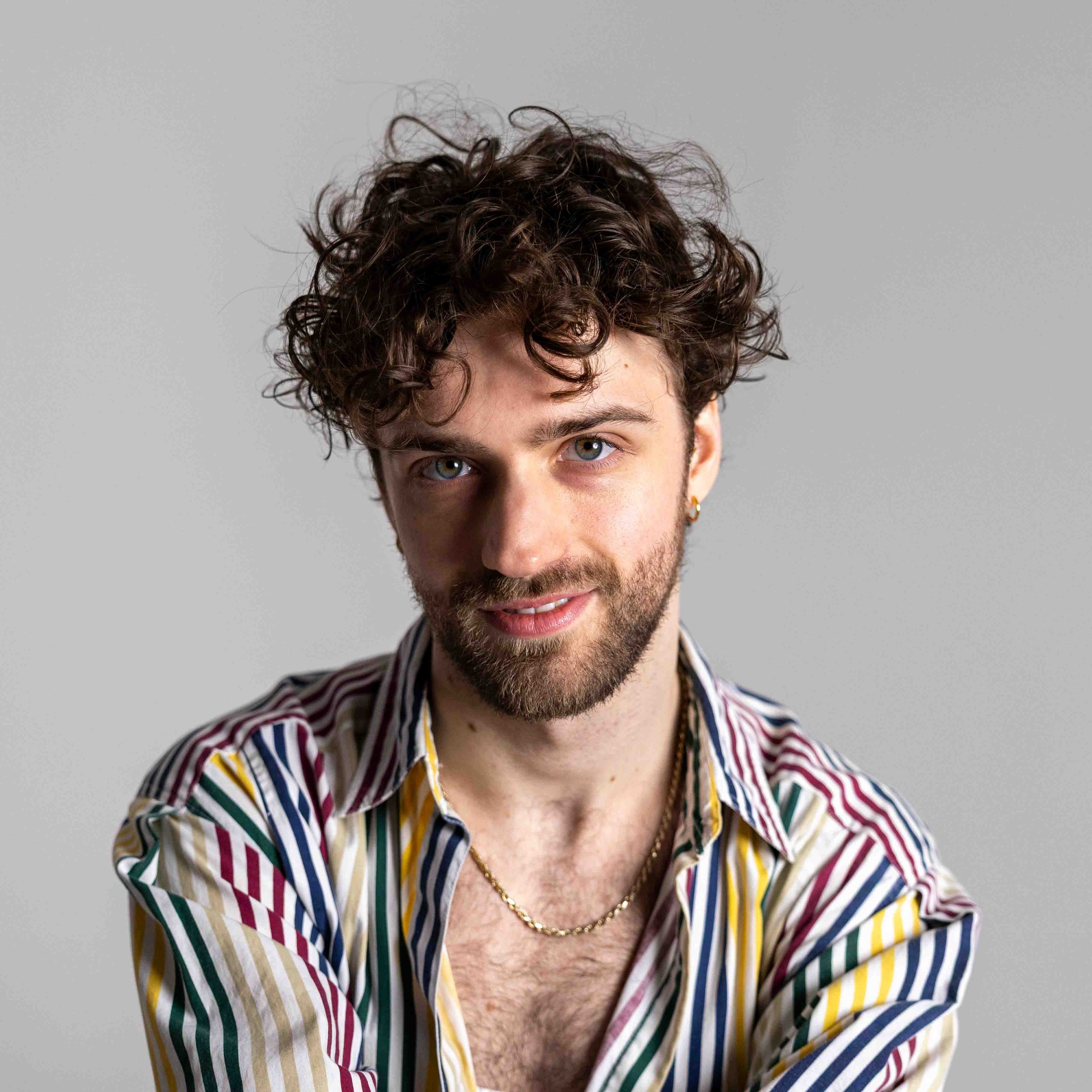 A man with curly brown hair, blue eyes, a beard, wearing a striped multicolor shirt, gold chain, and earring, smiling at the camera against a gray background.