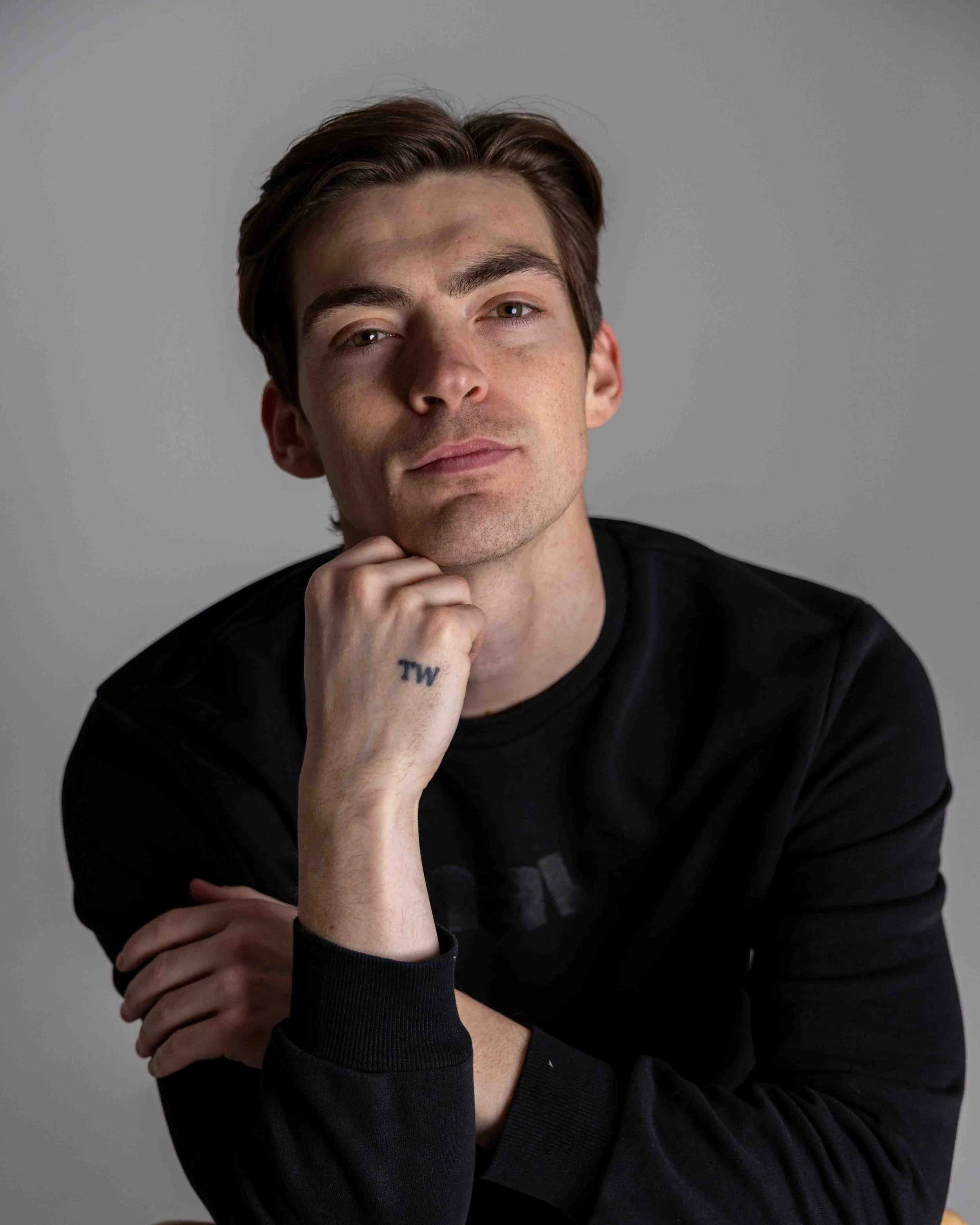 Young man with brown hair wearing a black long-sleeve shirt, sitting against a gray background, resting his chin on his hand, with a small tattoo 'TW' on his wrist.