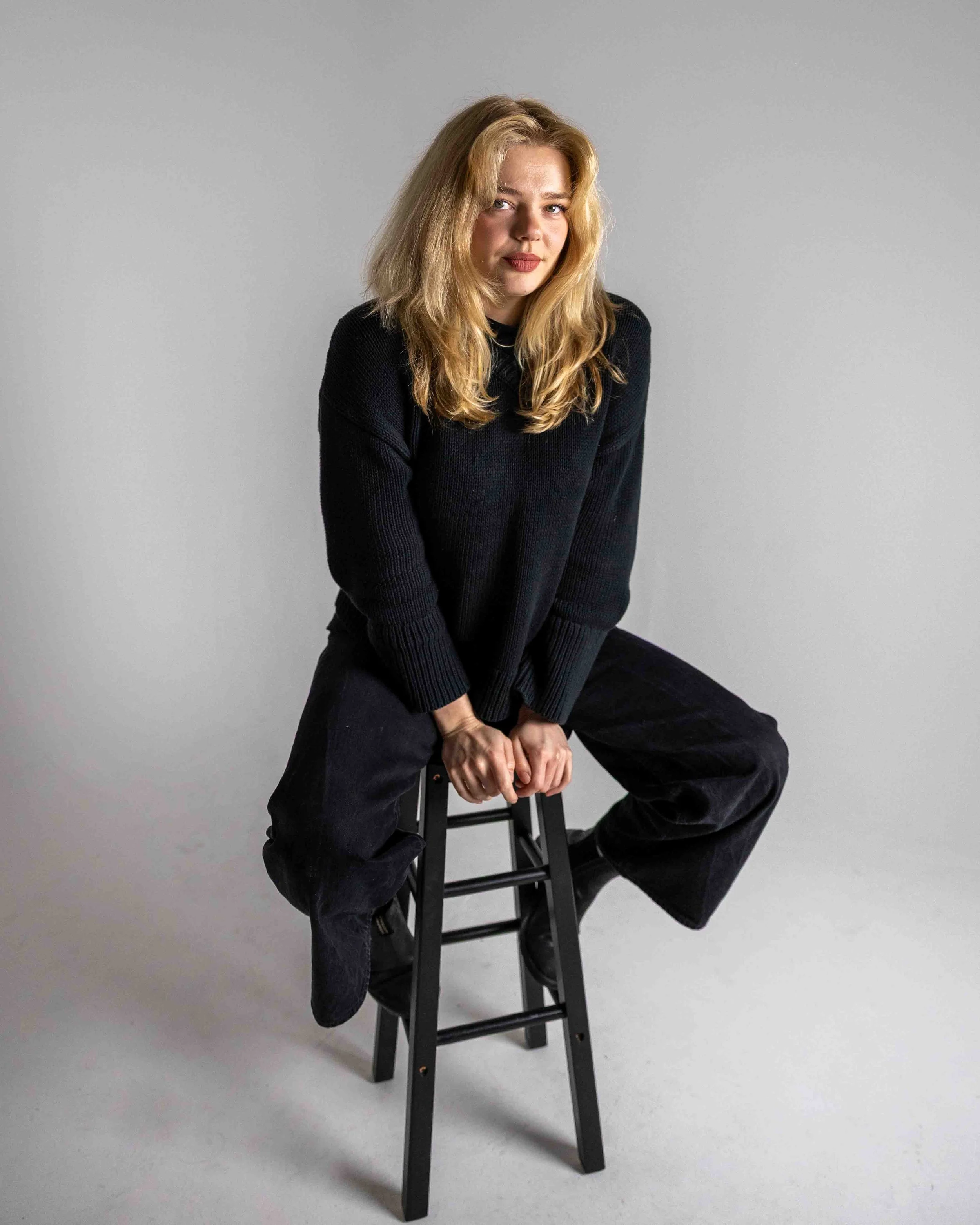 A woman with blonde hair and a black sweater sitting on a black step stool against a plain gray background.