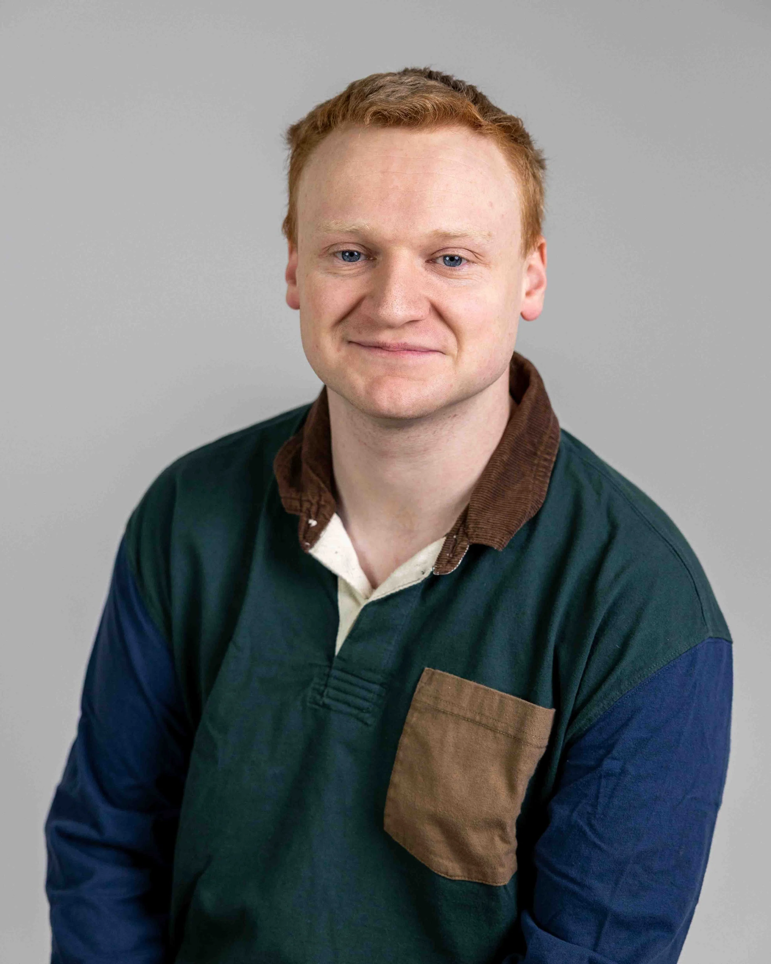 A young man with short red hair and blue eyes smiling, wearing a multicolored long-sleeve shirt with a brown collar and pocket, against a plain gray background.
