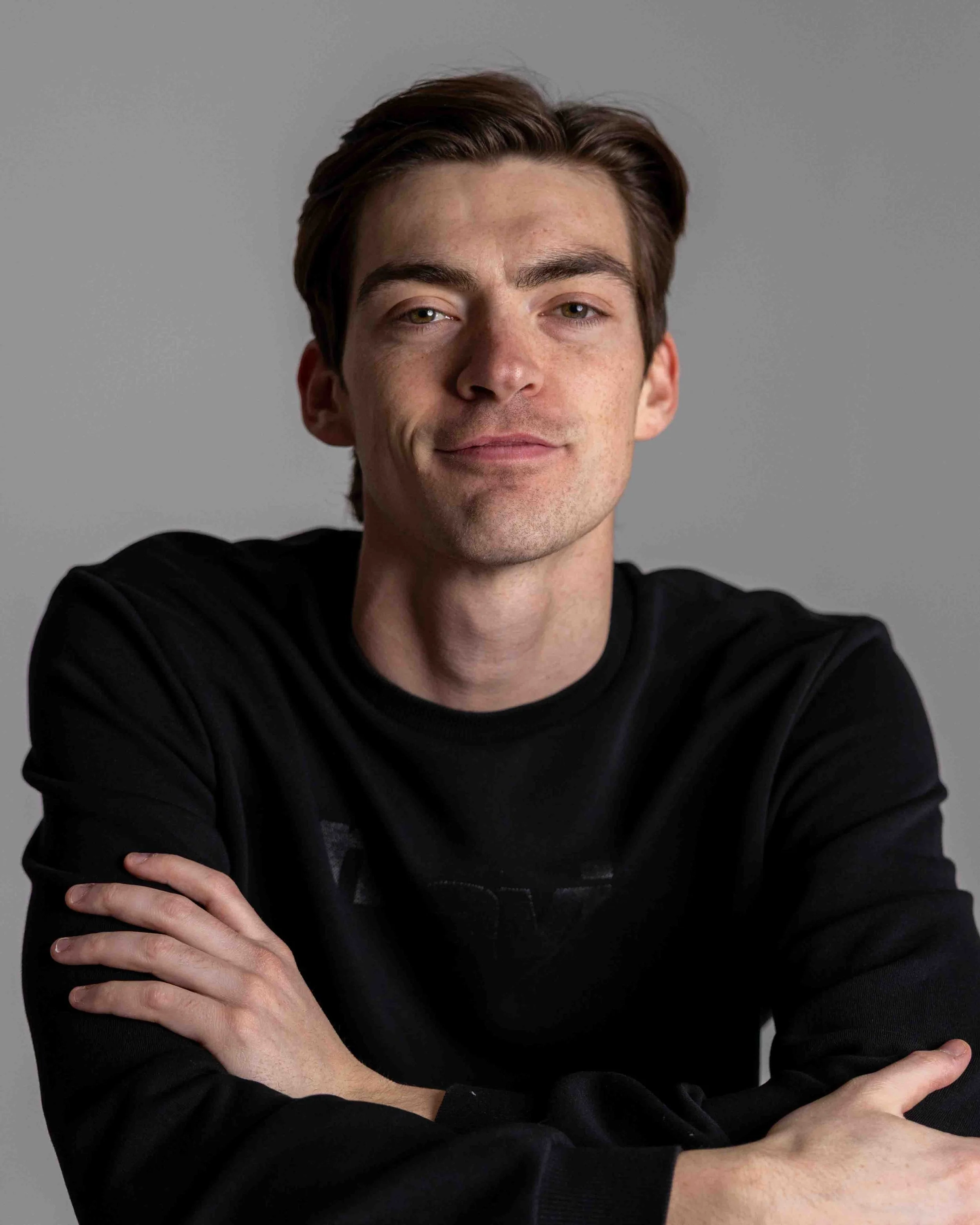 A young man with brown hair, fair skin, and green eyes, wearing a black long-sleeved shirt, looking at the camera with a slight smile, set against a plain gray background.