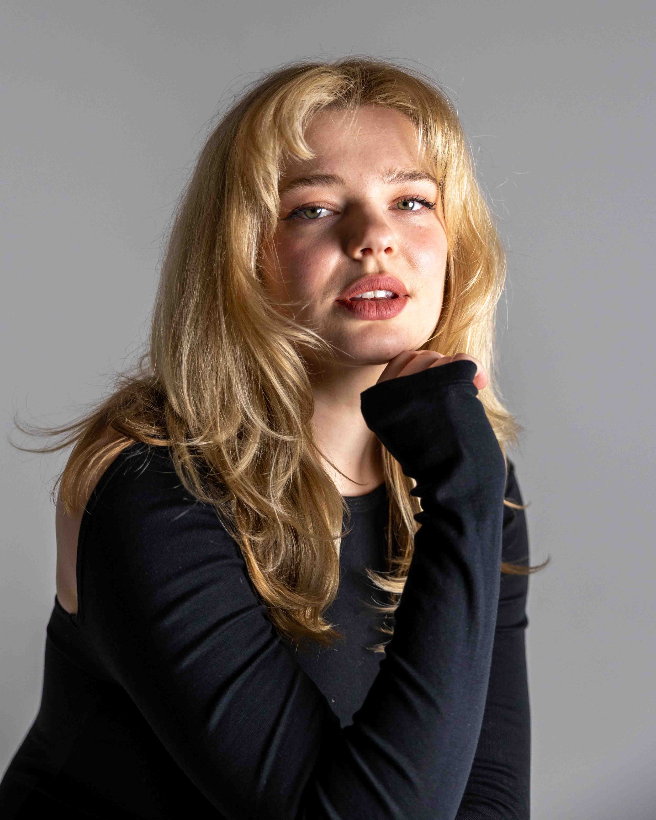 A young woman with blonde hair, wearing a black long sleeve top, posing in front of a plain gray background.