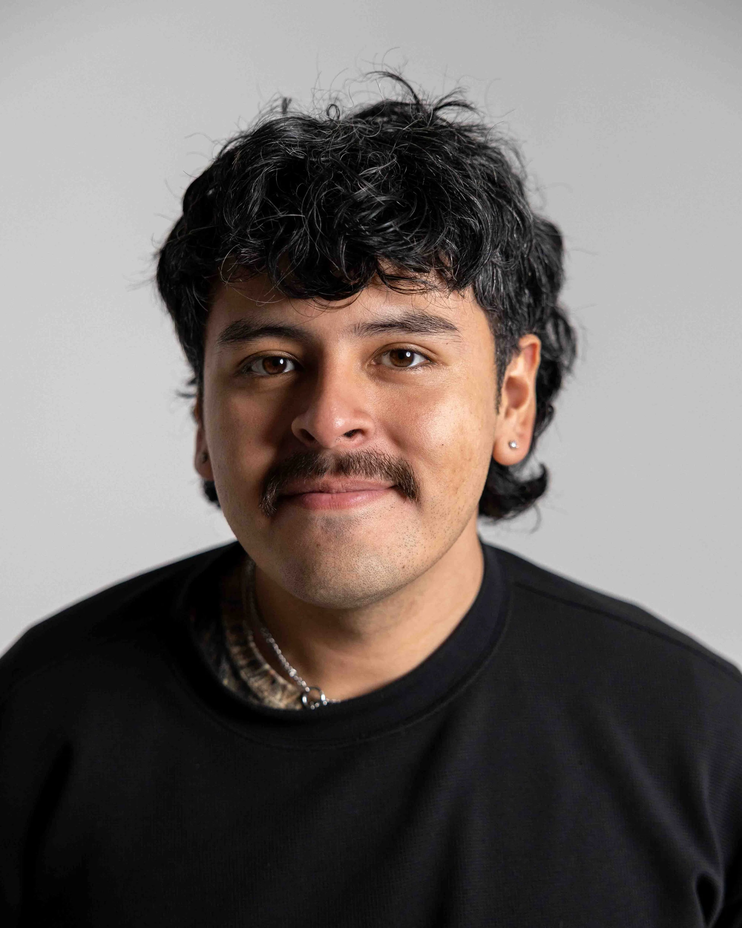 Portrait of a young man with dark curly hair, a mustache, wearing a black shirt and a necklace, against a light gray background.