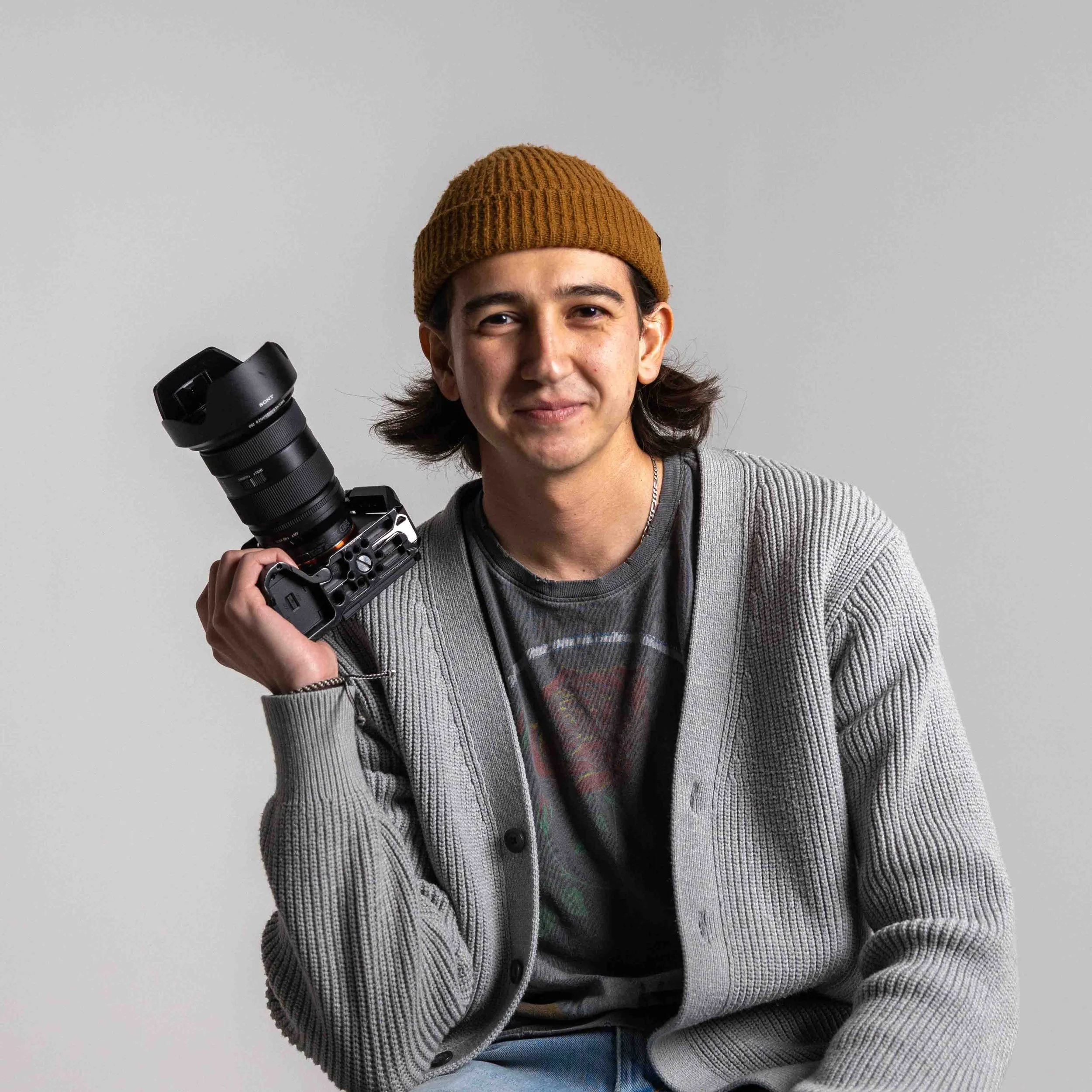 Young man with long hair, wearing a brown beanie, gray cardigan, and a graphic T-shirt, holding a camera with a large lens, smiling against a plain gray background.