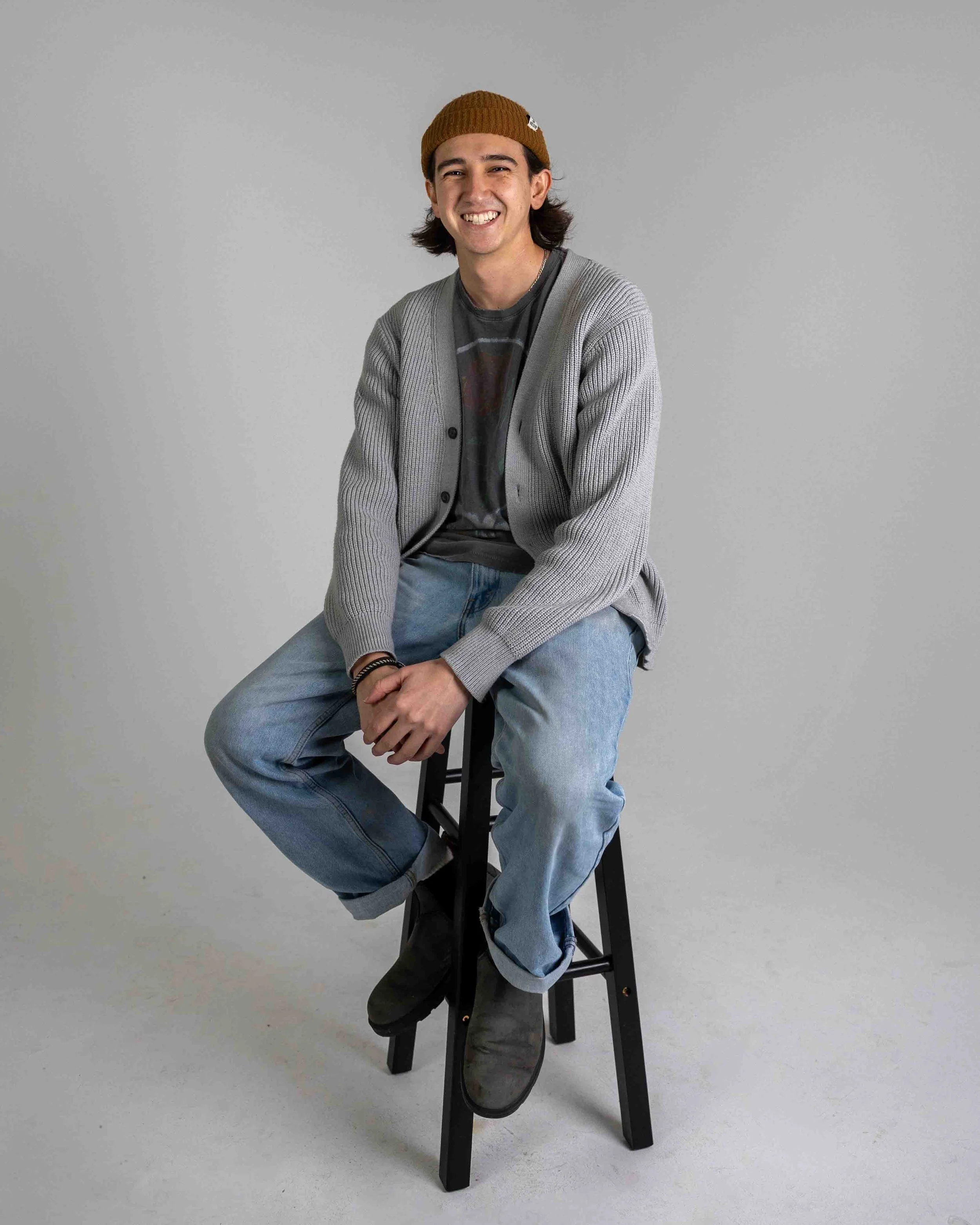 Young man sitting on a tall black stool, smiling, wearing a brown knit beanie, gray cardigan, graphic t-shirt, light blue jeans, and gray boots, against a plain light gray background.