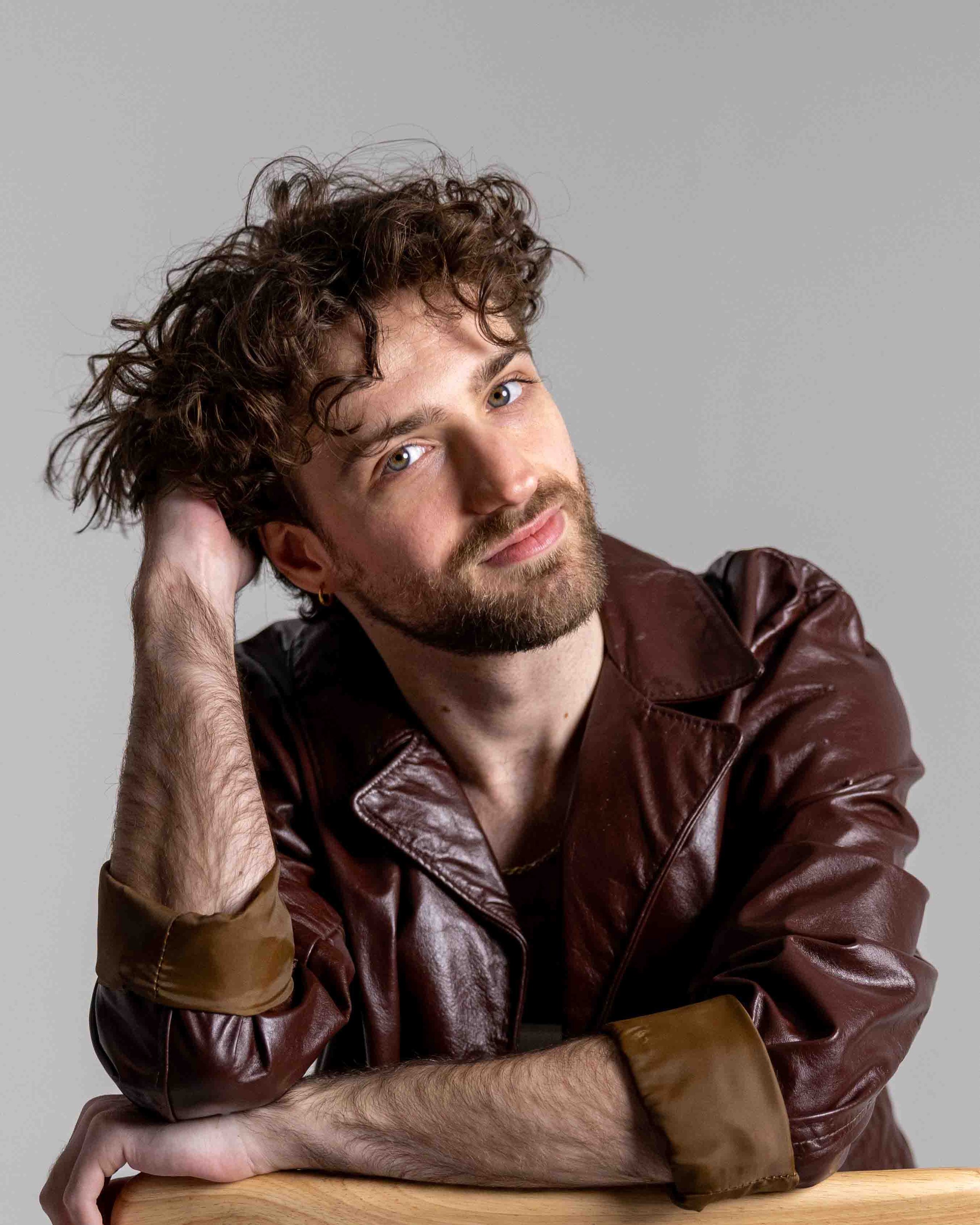 Portrait of a young man with curly hair and a beard, wearing a brown leather jacket and a gold earring, sitting at a wooden surface against a plain background.
