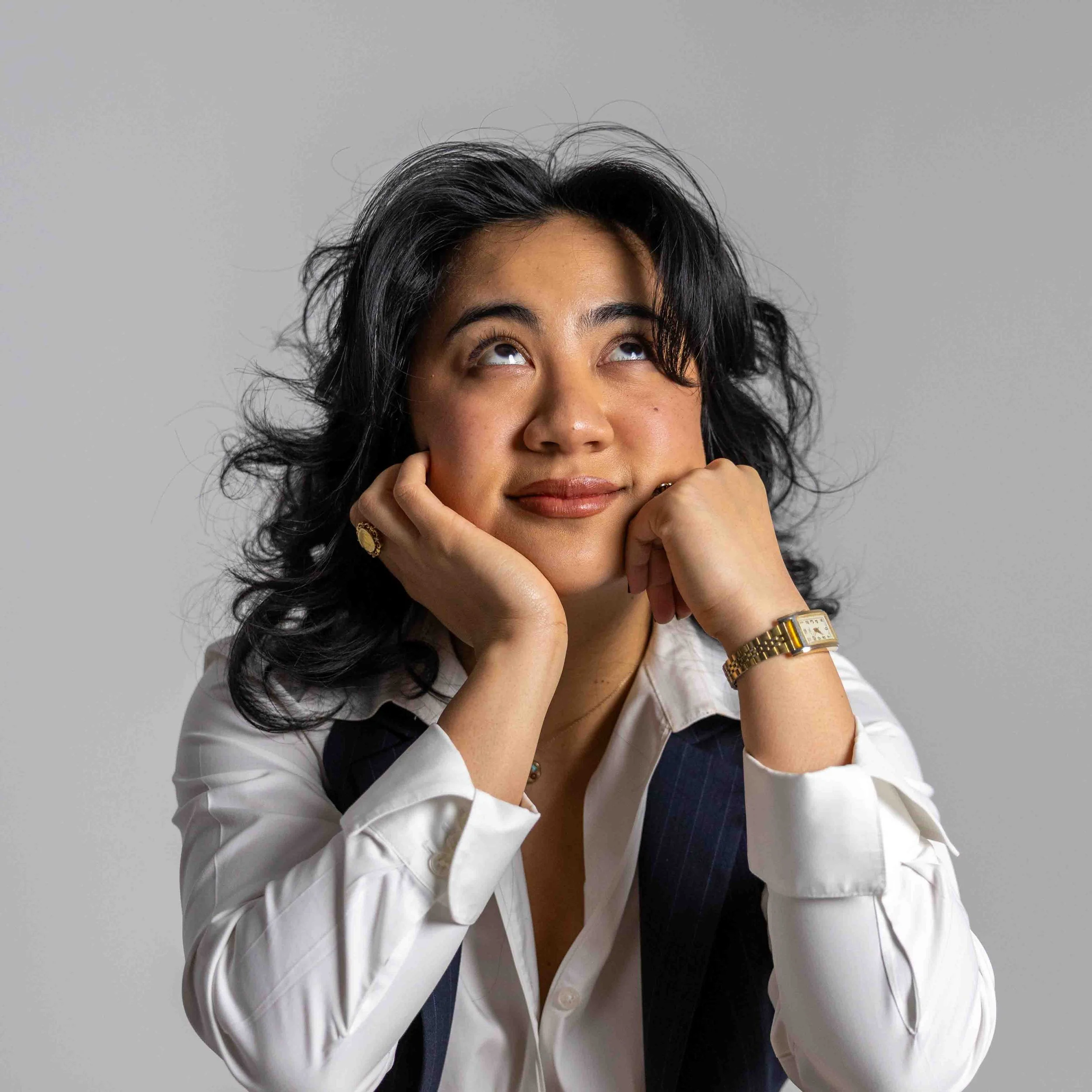Woman with dark curly hair wearing a white shirt, resting her chin on her hands, looking upwards with a thoughtful expression.