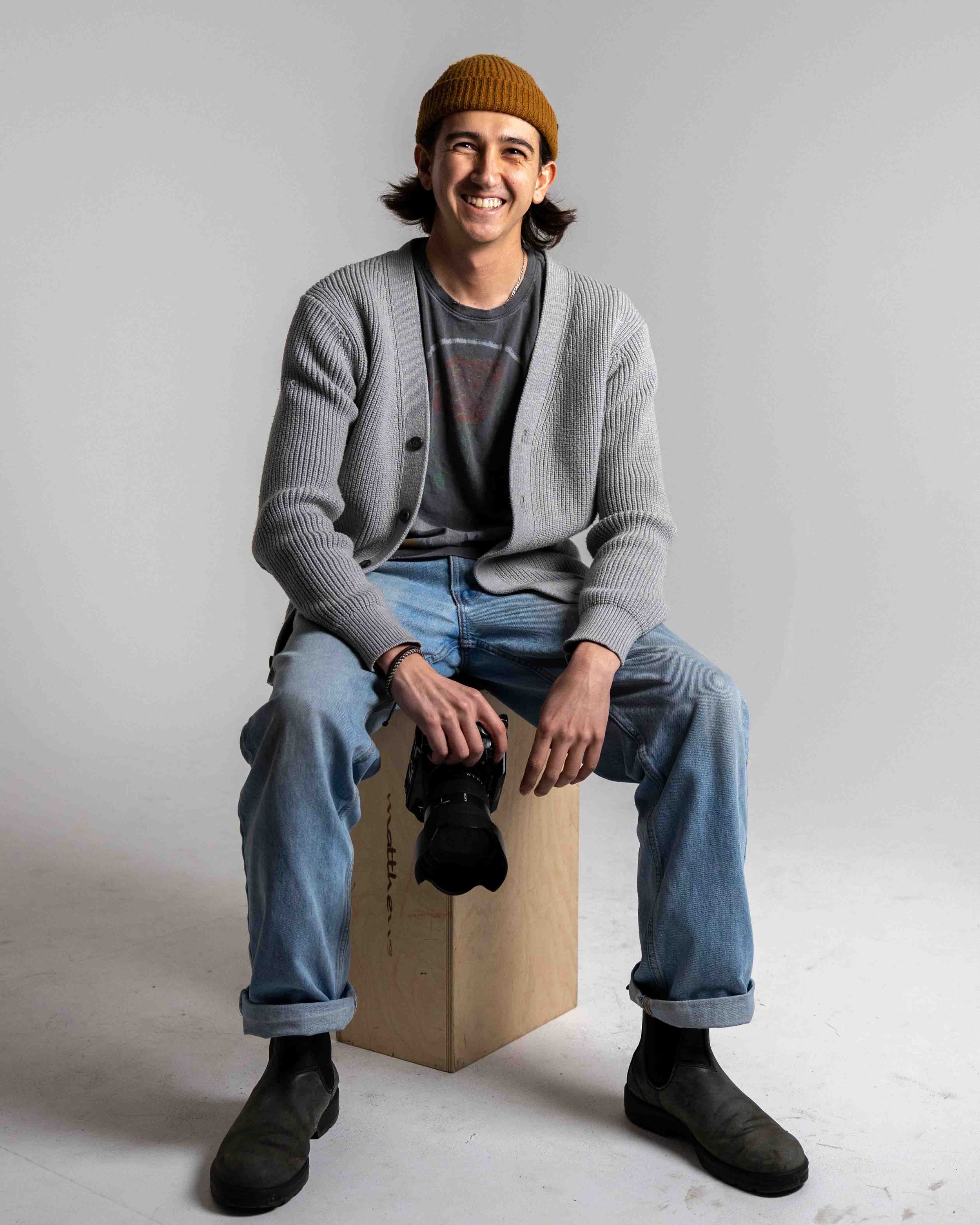 A young man sitting on a wooden box, holding a camera, smiling, wearing a yellow beanie, gray cardigan, t-shirt, jeans, and black boots against a plain gray background.