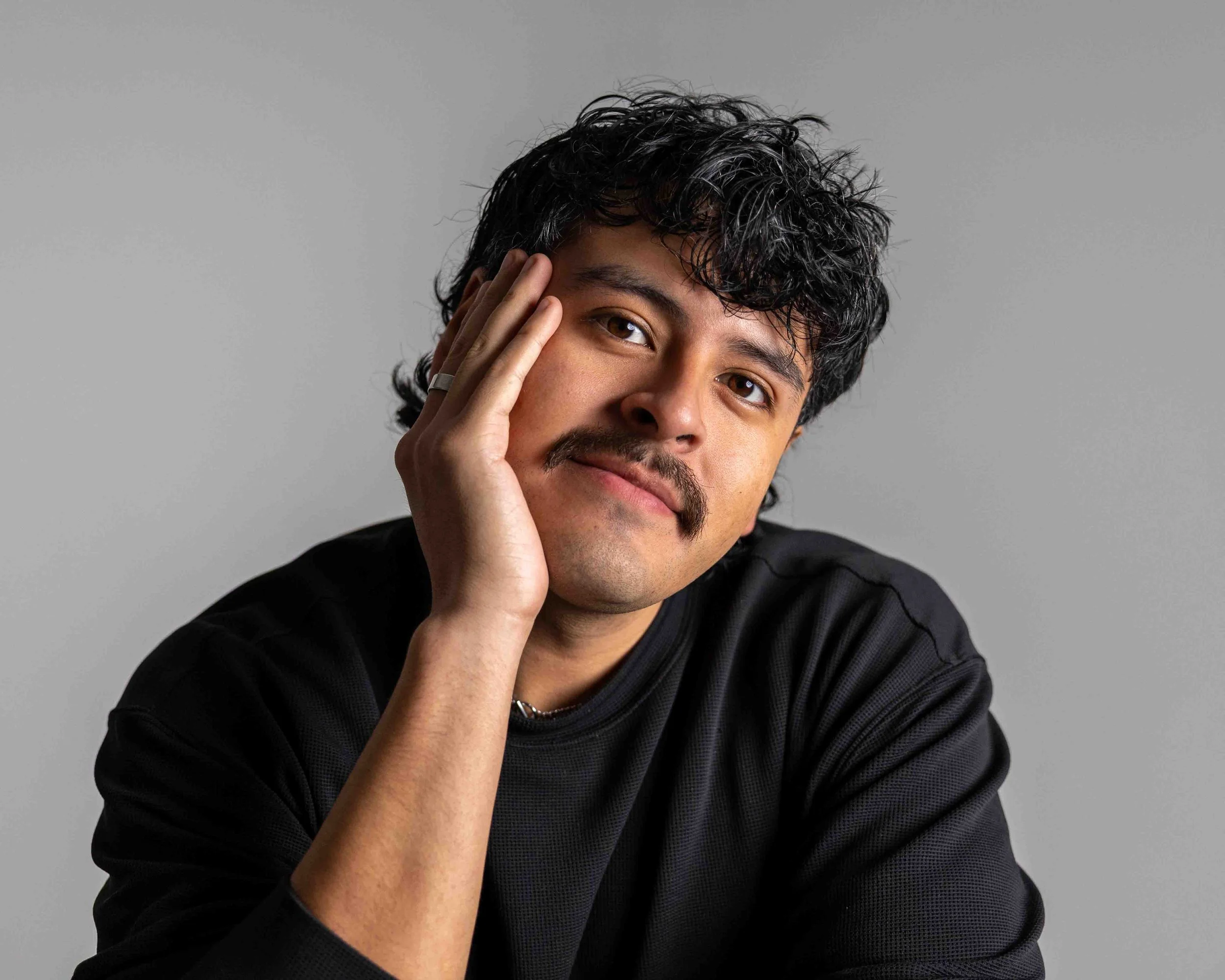A man with dark curly hair and a mustache resting his face on his hand, wearing a black shirt, against a plain gray background.