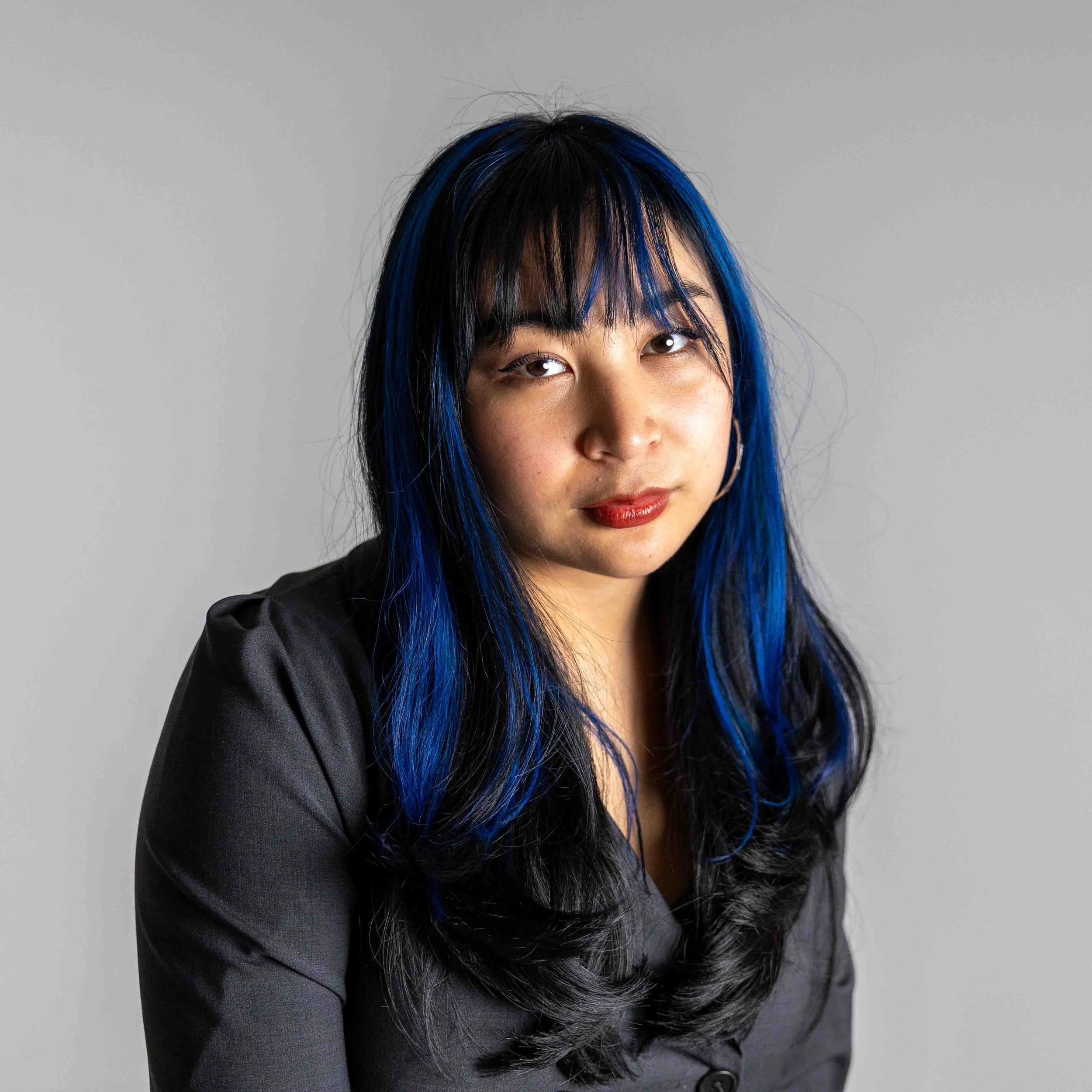 Young woman with black and blue hair, wearing a black blazer, looking at the camera against a plain gray background.