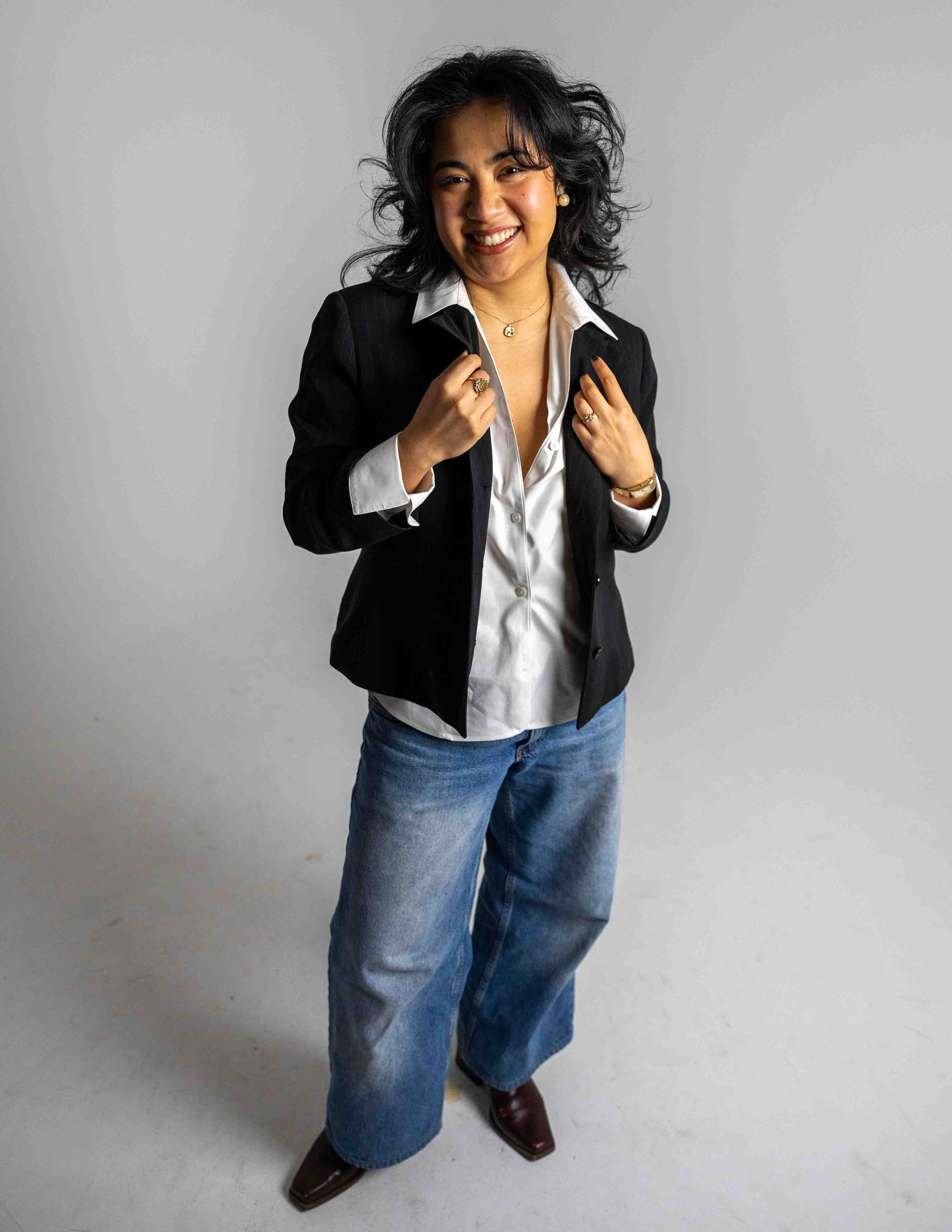A woman with black curly hair, smiling and dressed in a black blazer, white shirt, and blue jeans, standing against a gray background.