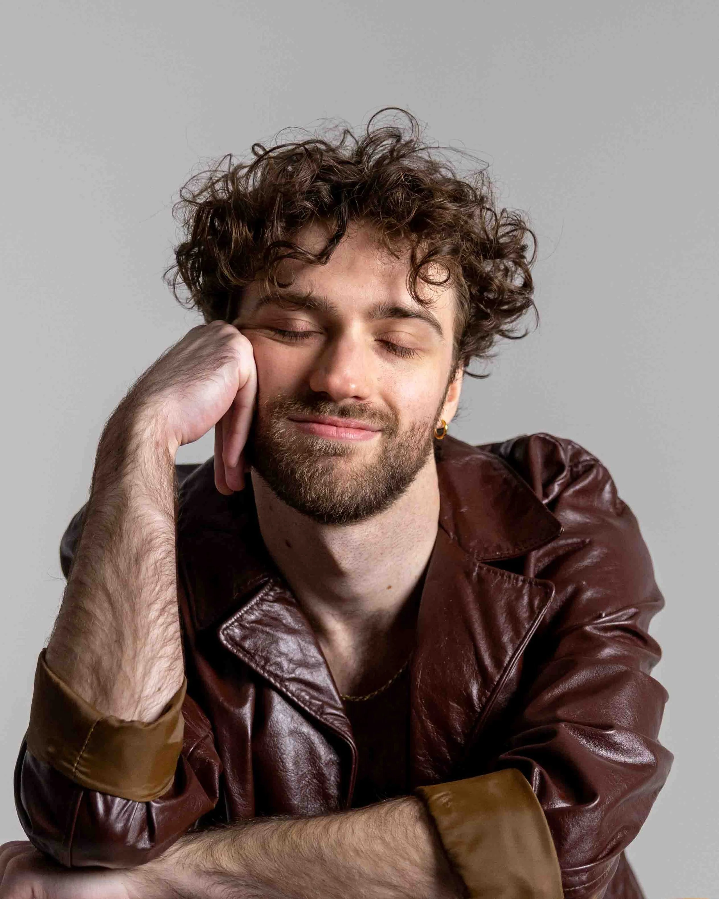 A man with curly hair and a beard, smiling with his eyes closed, resting his head on his left hand, wearing a brown leather jacket and a gold earring, against a gray background.