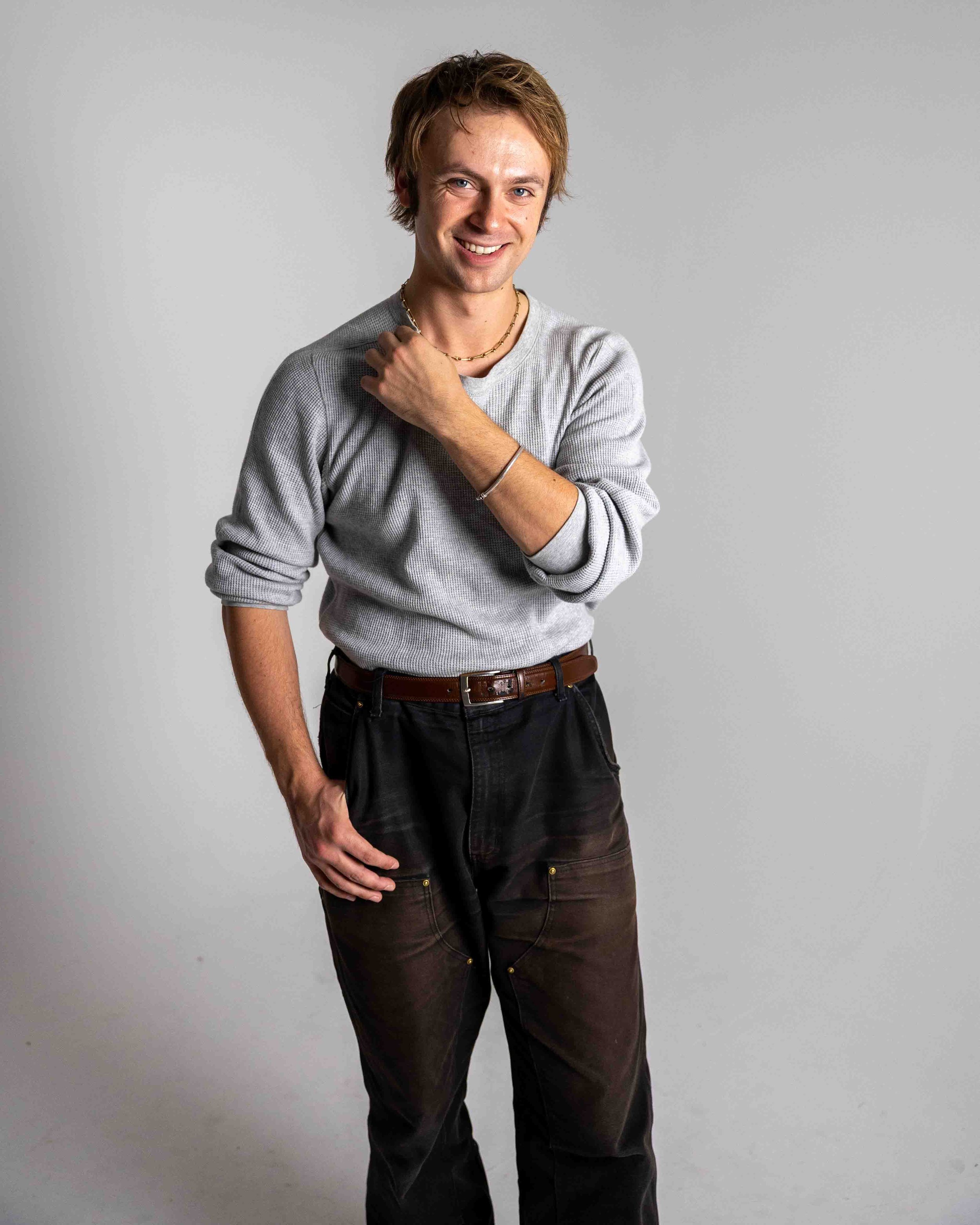 A young man with ginger hair wearing a gray long-sleeve shirt, black pants, and accessories, standing against a plain light gray background, smiling, with one hand on his chest and the other resting on his hip.