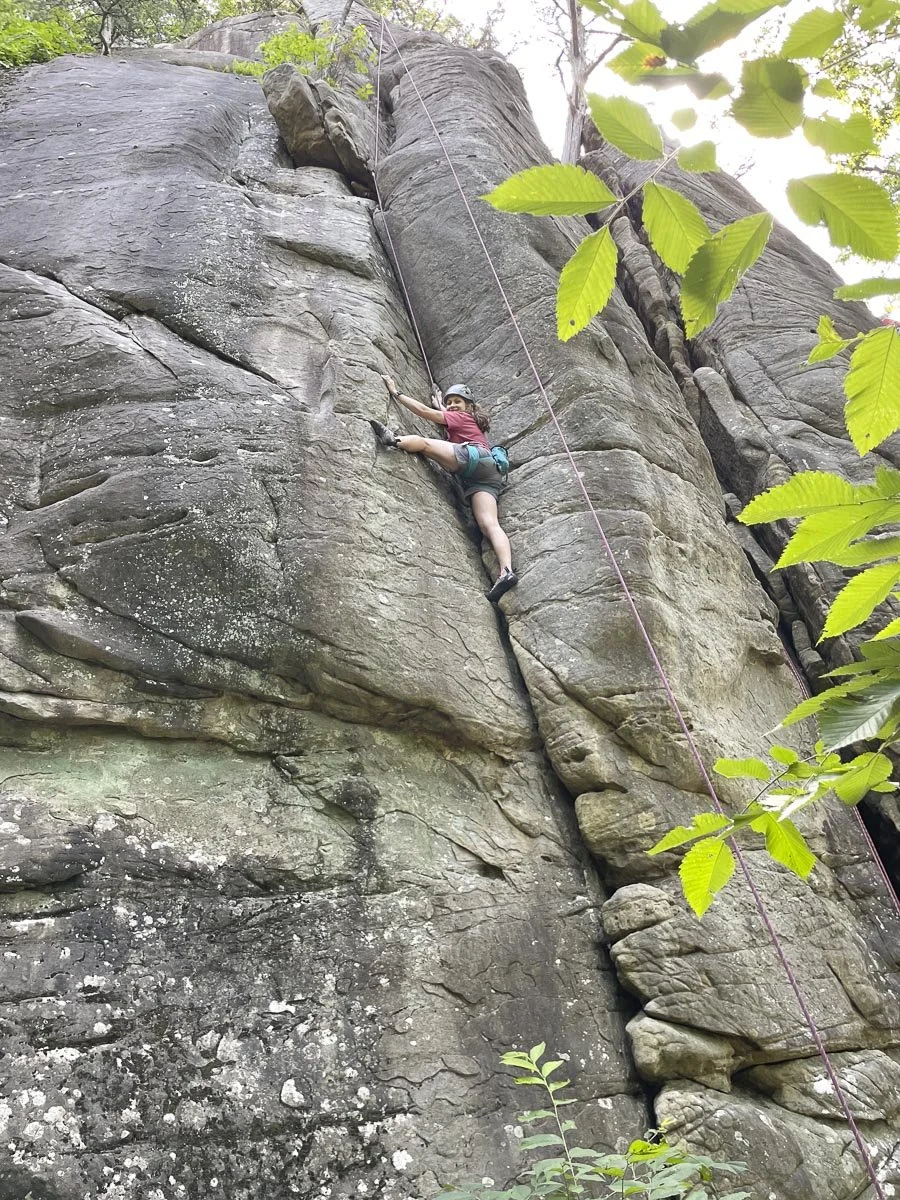 A woman rock climbing on a tall, rugged cliff face with green foliage around.