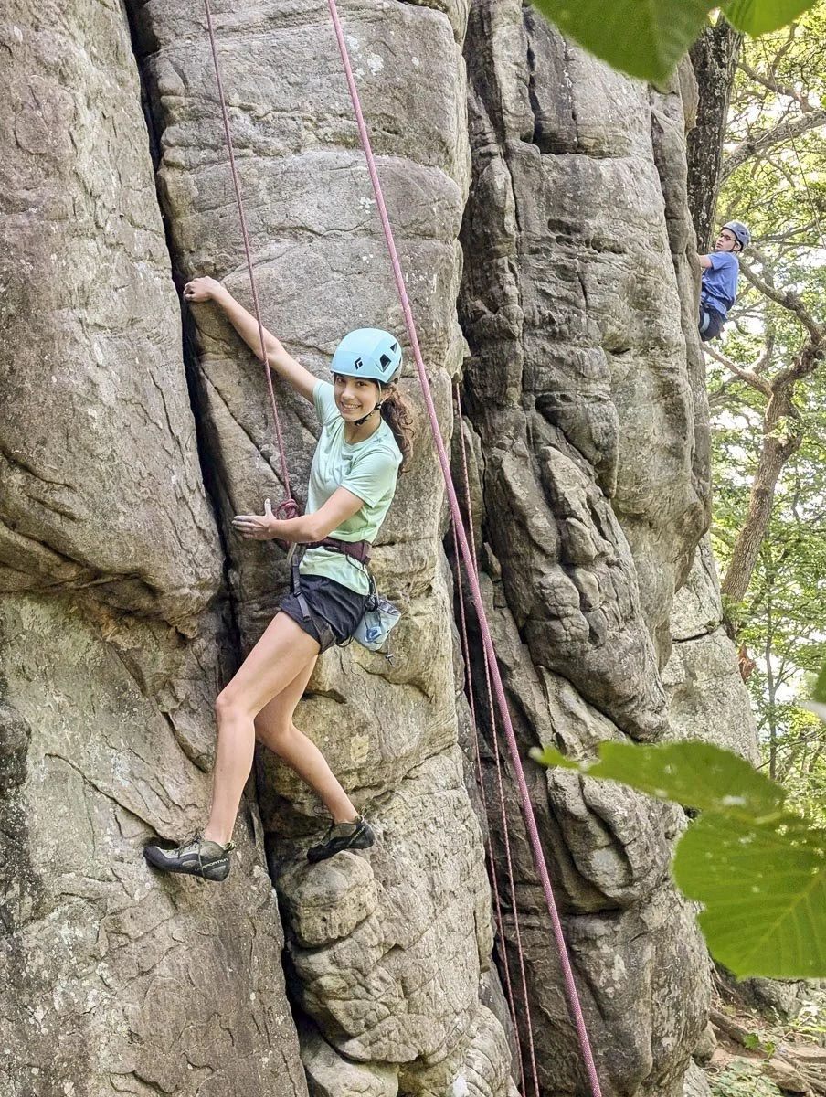 A woman rock climbing on a vertical rock formation with a safety harness and helmet, smiling at the camera. A man is climbing higher on the cliff in the background.