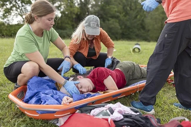 Two women load a female patient with a head injury into a bright orange medical litter in a grassy field. The women on the left gestures with her hand and is speaking. A third person's hands and legs border the right side of the image.