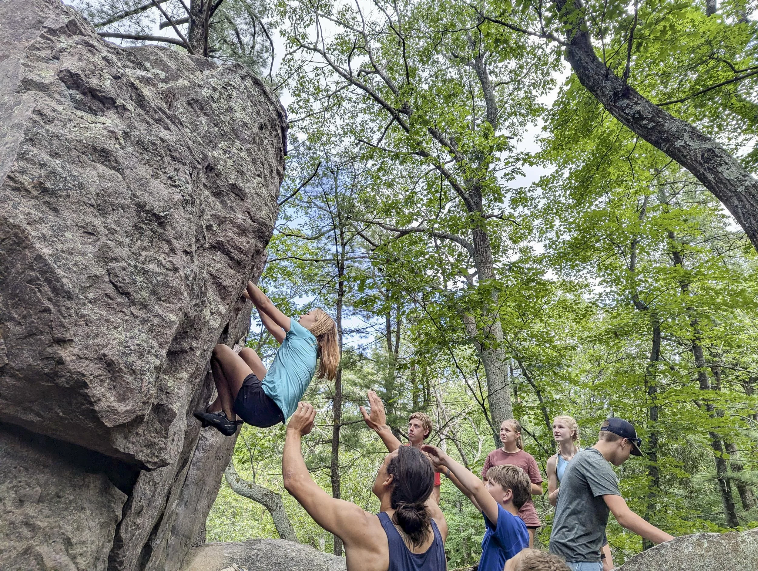 Group of people assisting girl bouldering outdoors in a wooded area