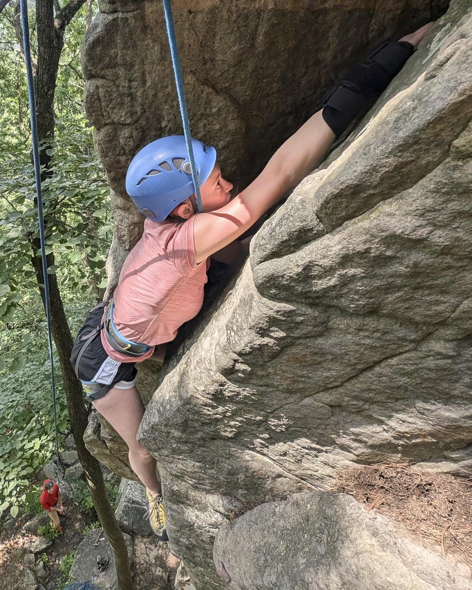 A woman wearing a blue helmet, pink shirt, and black shorts is bouldering on a large rock formation outdoors, with a person in a red shirt and helmet at the bottom.