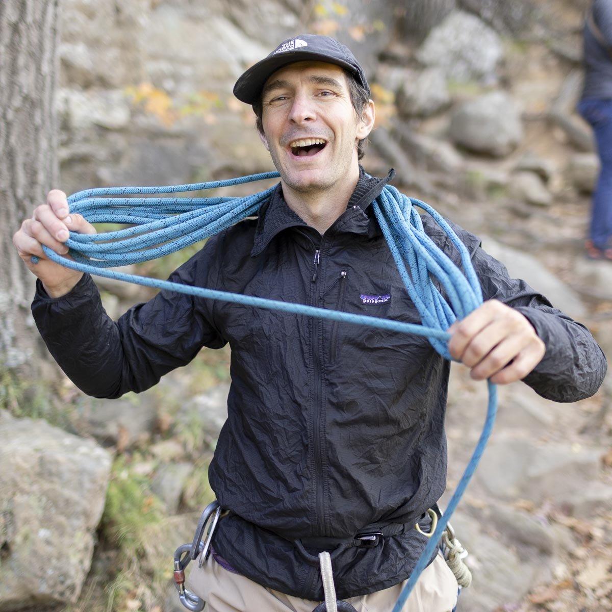 A man smiling outdoors, wearing a black jacket and carrying blue climbing ropes on his shoulder.