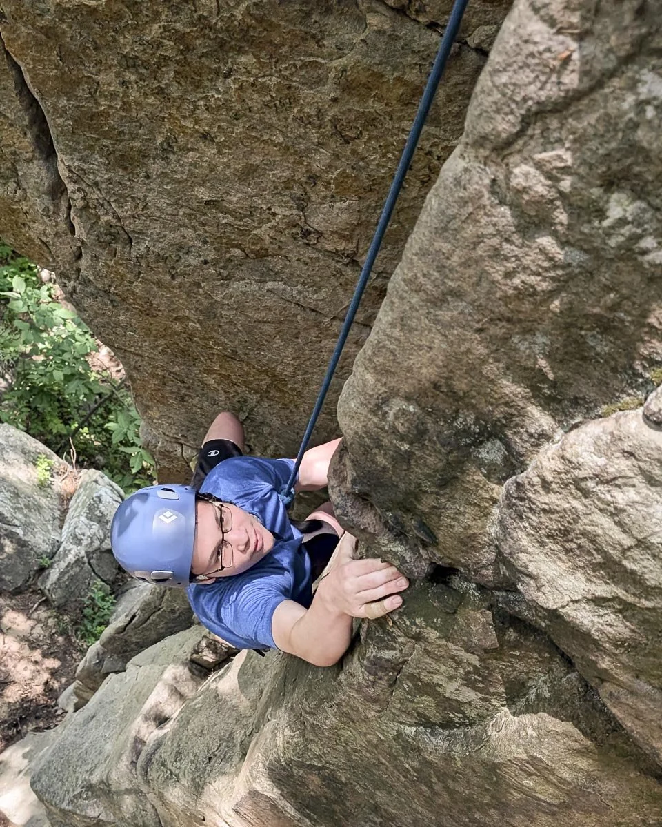 A person rock climbing, wearing a helmet and glasses, gripping a rocky surface with hands, and secured with a climbing harness and rope.