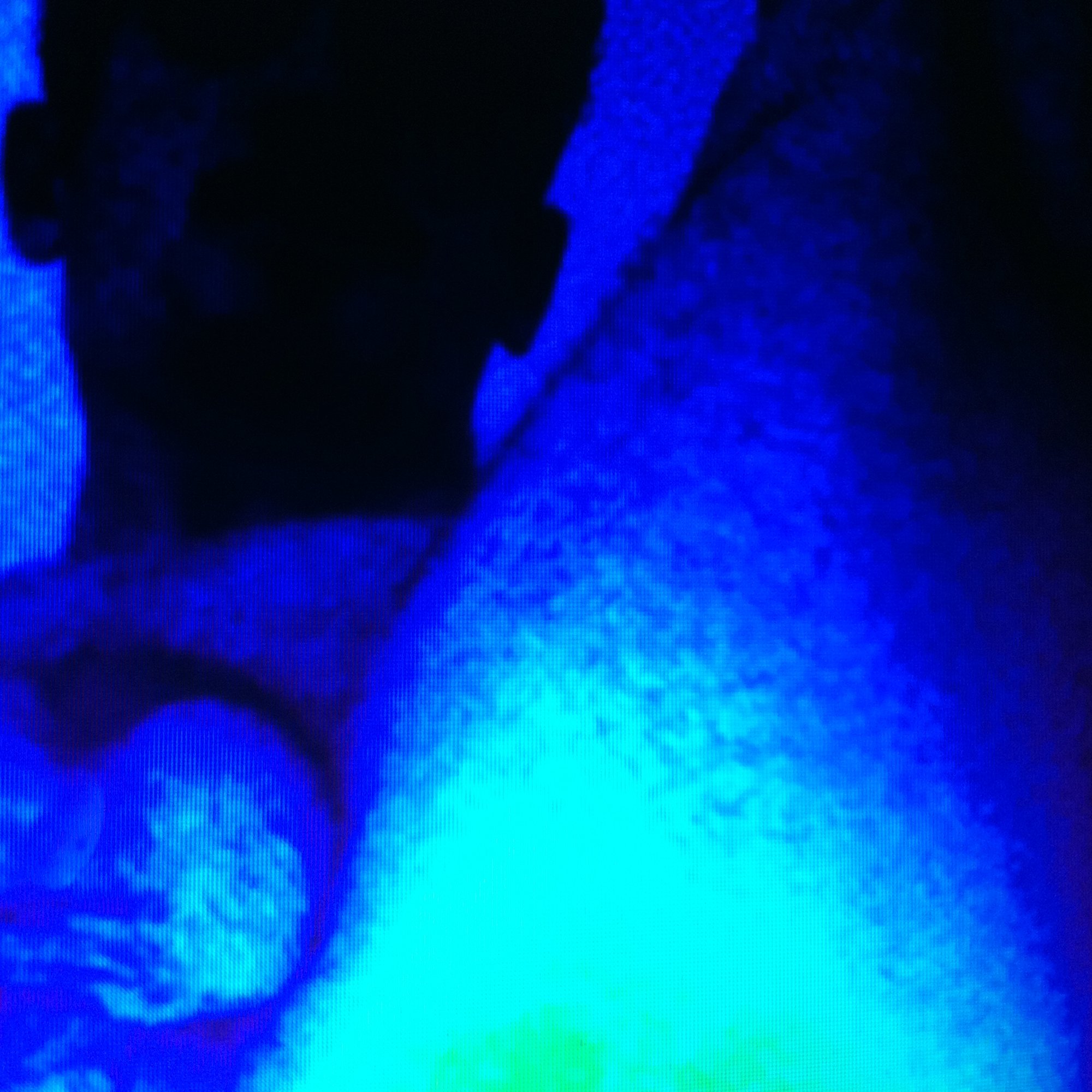 Close-up of a person's face in blue lighting, wearing glasses, with their hand near their ear.