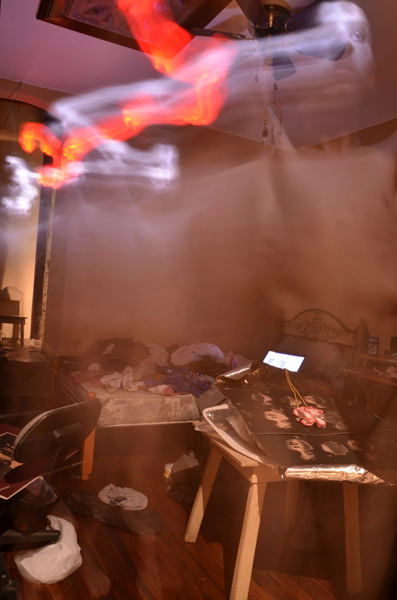 A bedroom filled with smoke and fire, with a ceiling fan, a mattress with clothes and items, and a black table covered with a spray-painted face design, a necklace, and other objects.