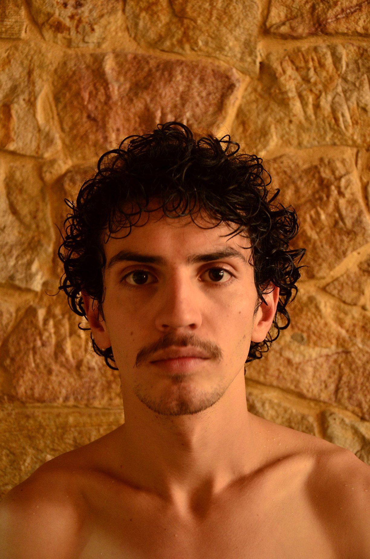 A shirtless young man with curly dark hair and a mustache stands in front of a textured stone wall.